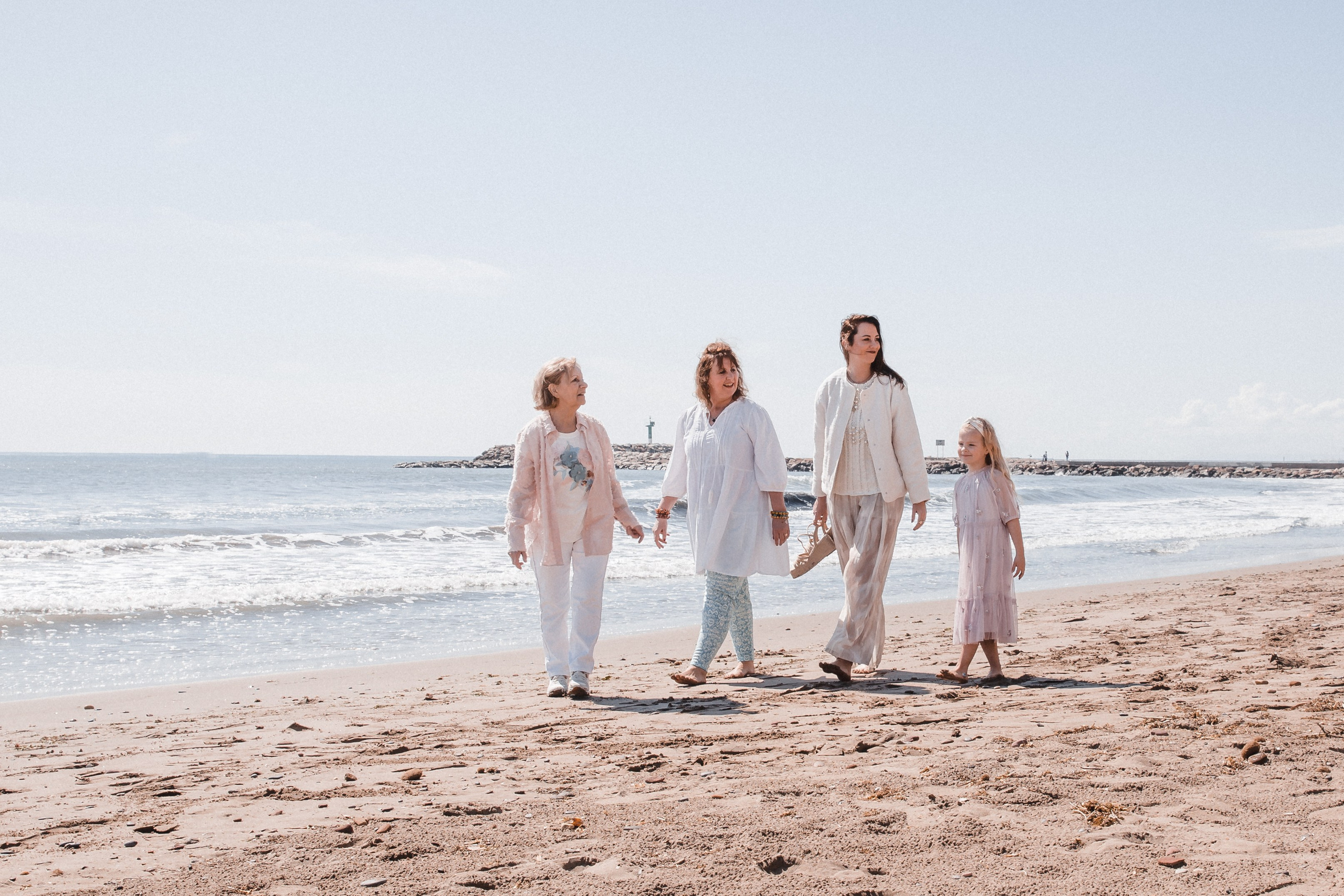 Cuatro generaciones de mujeres caminando de la mano por una playa tranquila en Valencia, España — retrato familiar conmovedor que refleja amor, legado y conexión junto al mar.