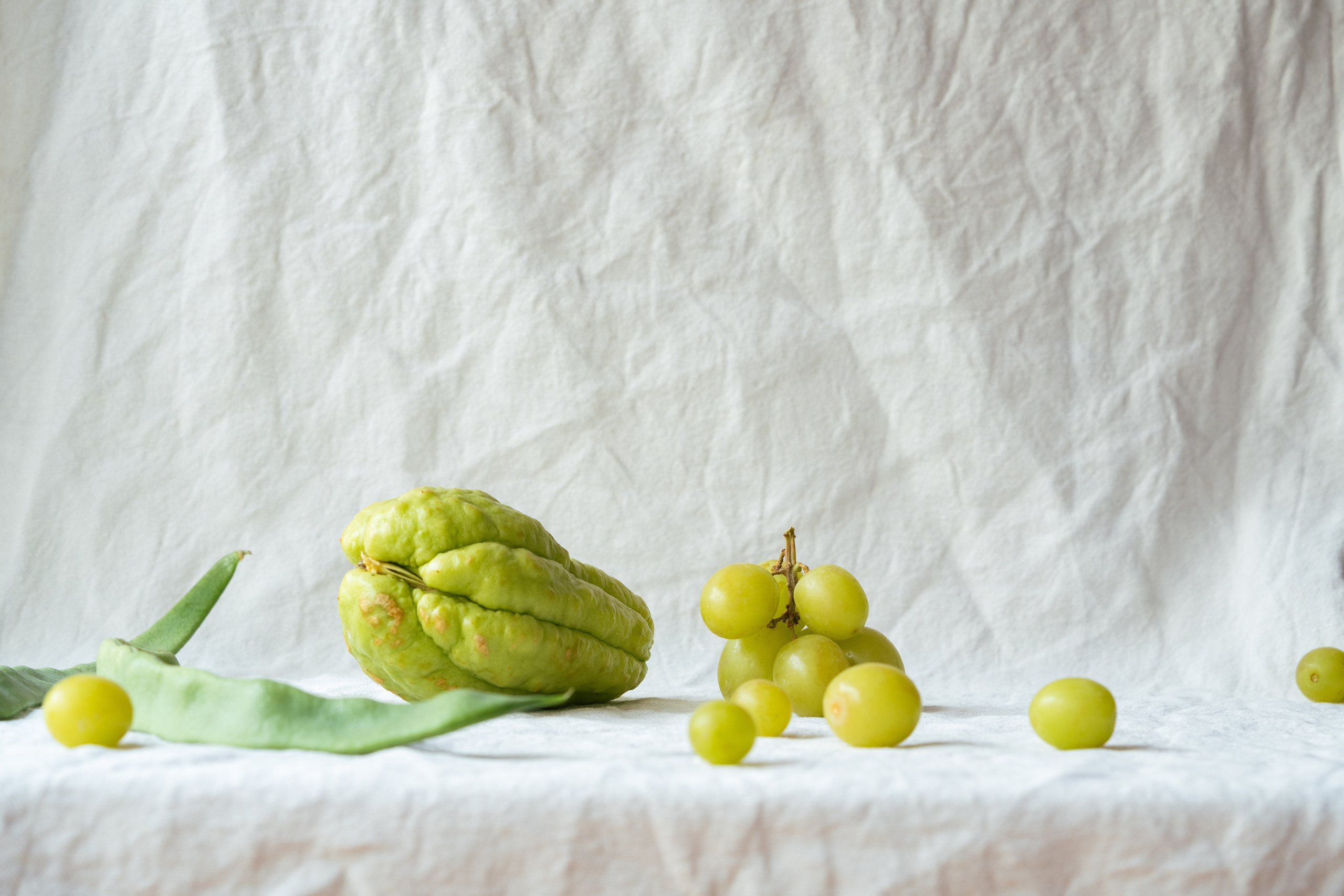 Fresh Produce Arrangement with Chayote and Grapes on White Fabric. Minimalist composition of fresh green chayote, grapes, and beans on a textured white backdrop.