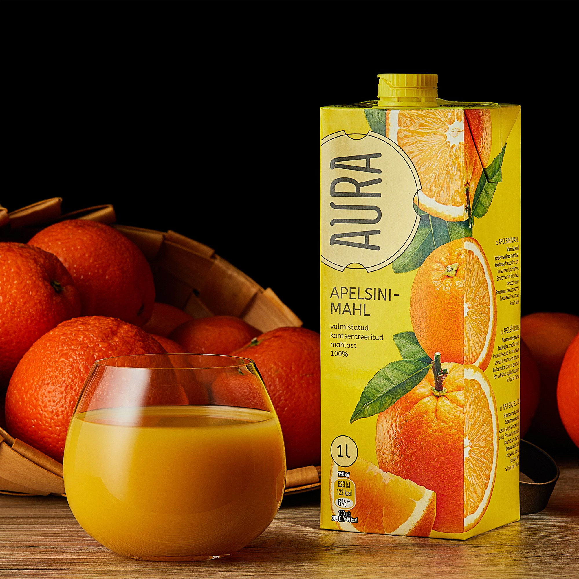 Photographer Roman Djuzev - The photograph shows AURA brand orange juice poured into a transparent glass, with a juice carton placed next to it. Behind the glass and the carton, part of a basket with oranges is visible, and the background is dark.