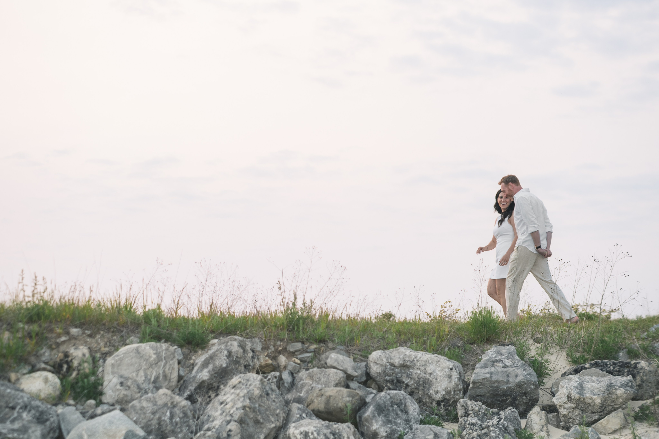 Engaged couple walking along a grassy hill during a romantic beachside photoshoot at sunset.