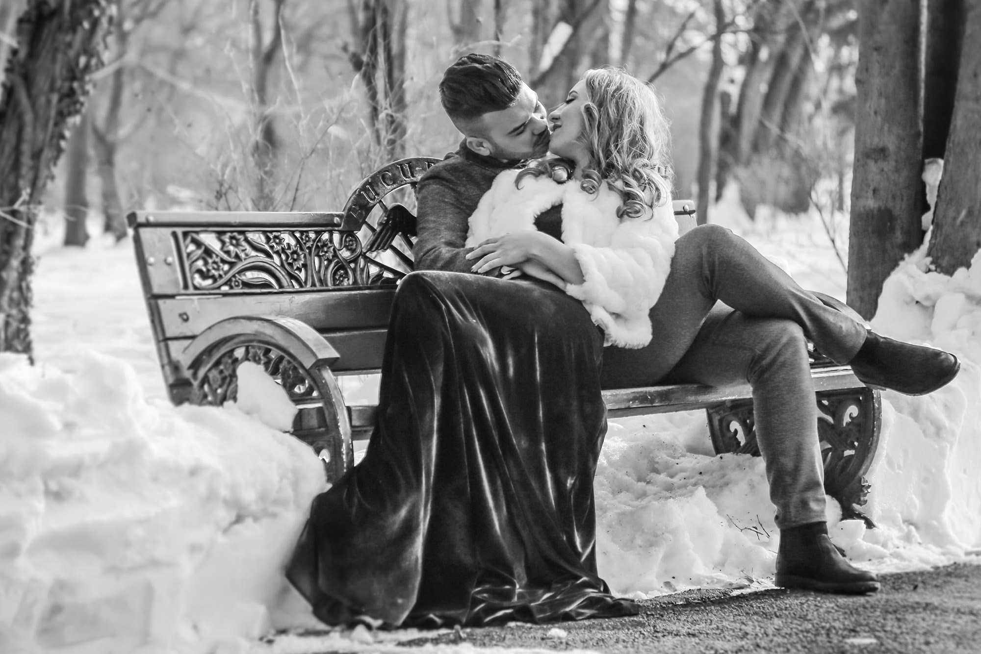 Romantic kiss between the newlyweds captured in black and white on a snowy park bench during their wedding photo shoot.