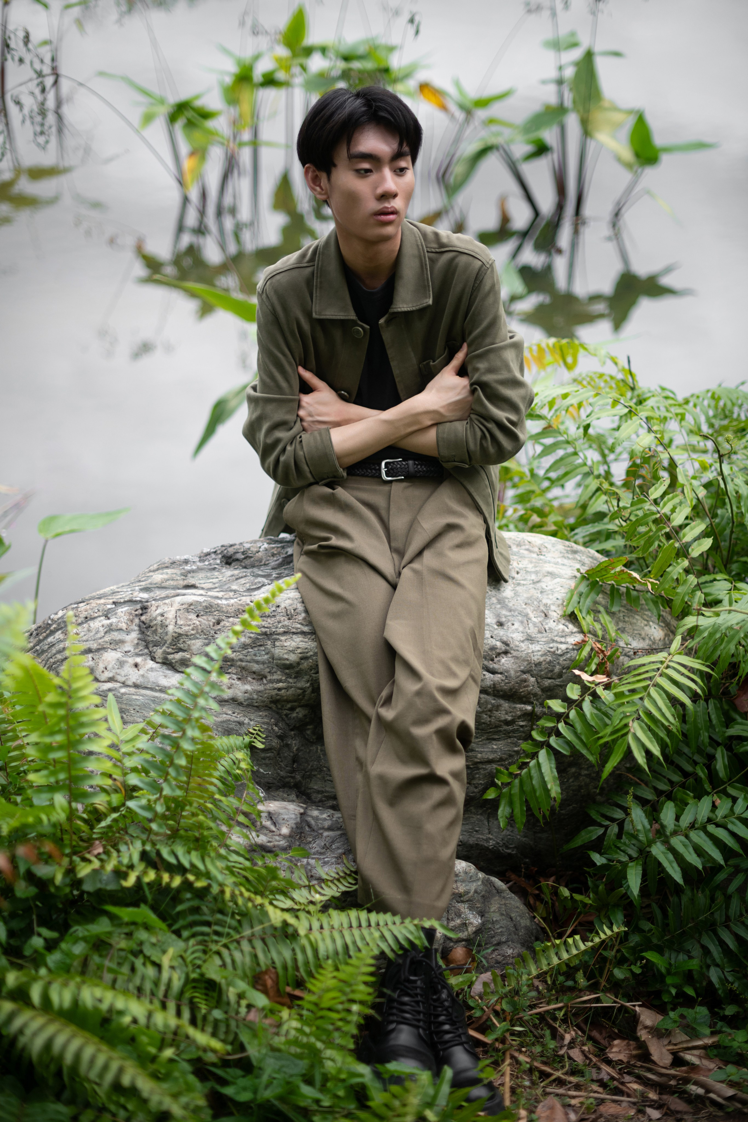 Editorial-style portrait of a young Asian man in olive jacket and khaki trousers sitting on a rock amidst ferns beside a reflective pond, Singapore.