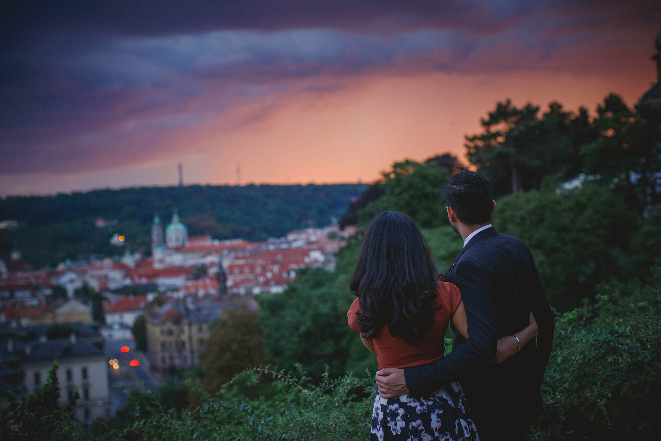 Stylish Indian couple embracing with vivid sky flaring above Prague skyline.