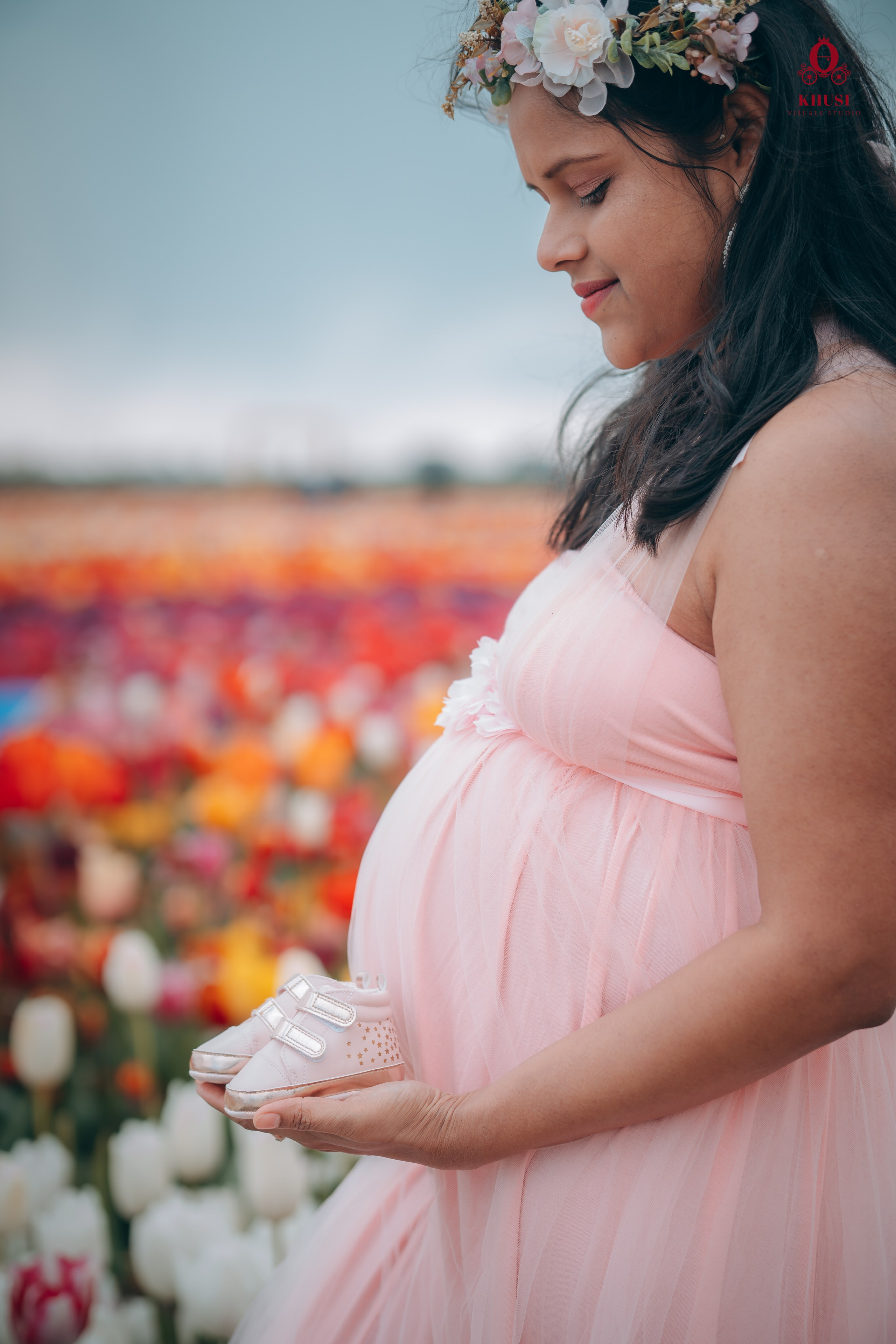 A pregnant mother is holding baby shoes and standing in a tulip flower field in netherlands