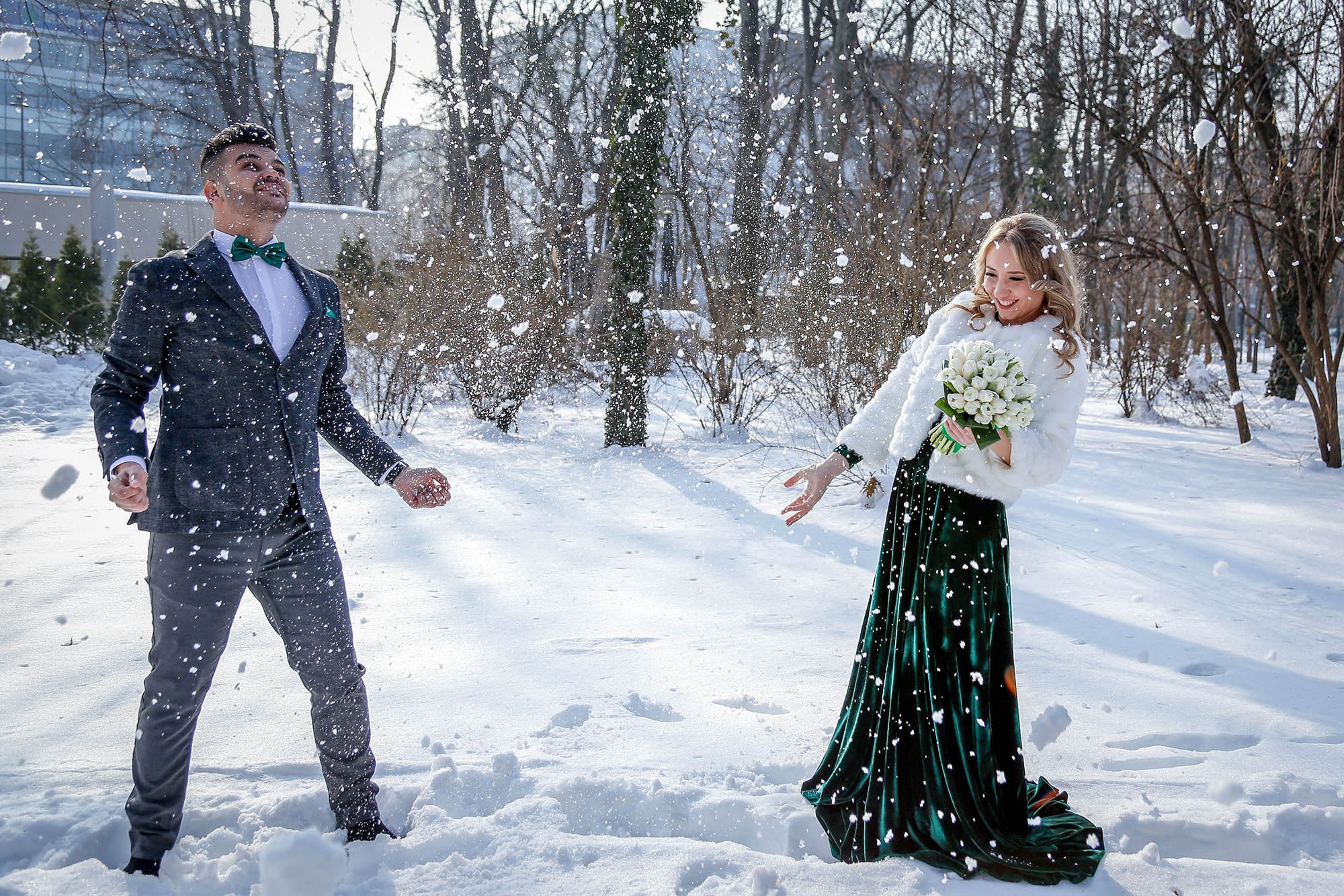 Happy newlyweds captured in a dynamic moment playing and throwing snow into the air during their park photo shoot.