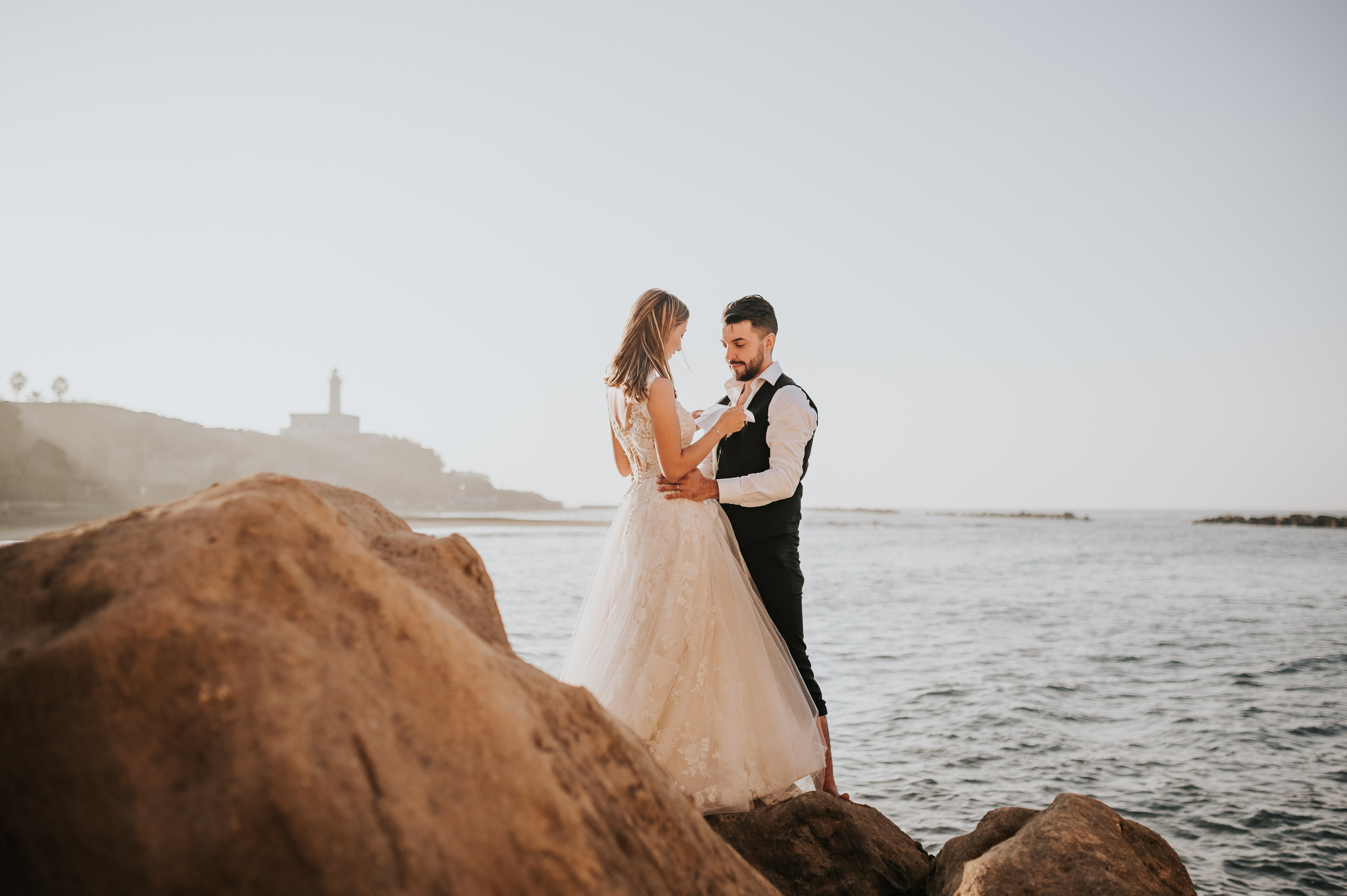 Coppia di neo-sposi in primo piano tra le rocce su una spiaggia al tramonto, con un faro sullo sfondo, immortalati da un fotografo di matrimonio.