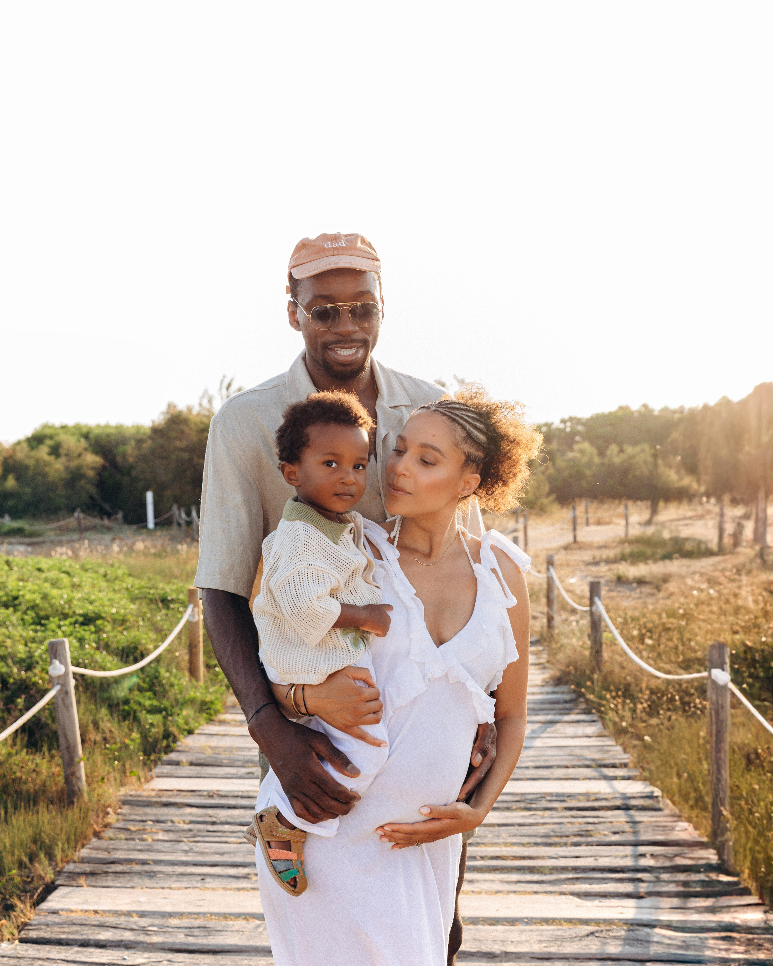 Sesión de embarazo familiar en Barcelona, España — padres alegres junto a su hijo pequeño abrazando esta nueva etapa con luz de sol sobre una pasarela. Inspiración perfecta para sesiones familiares de maternidad en Barcelona y toda España.