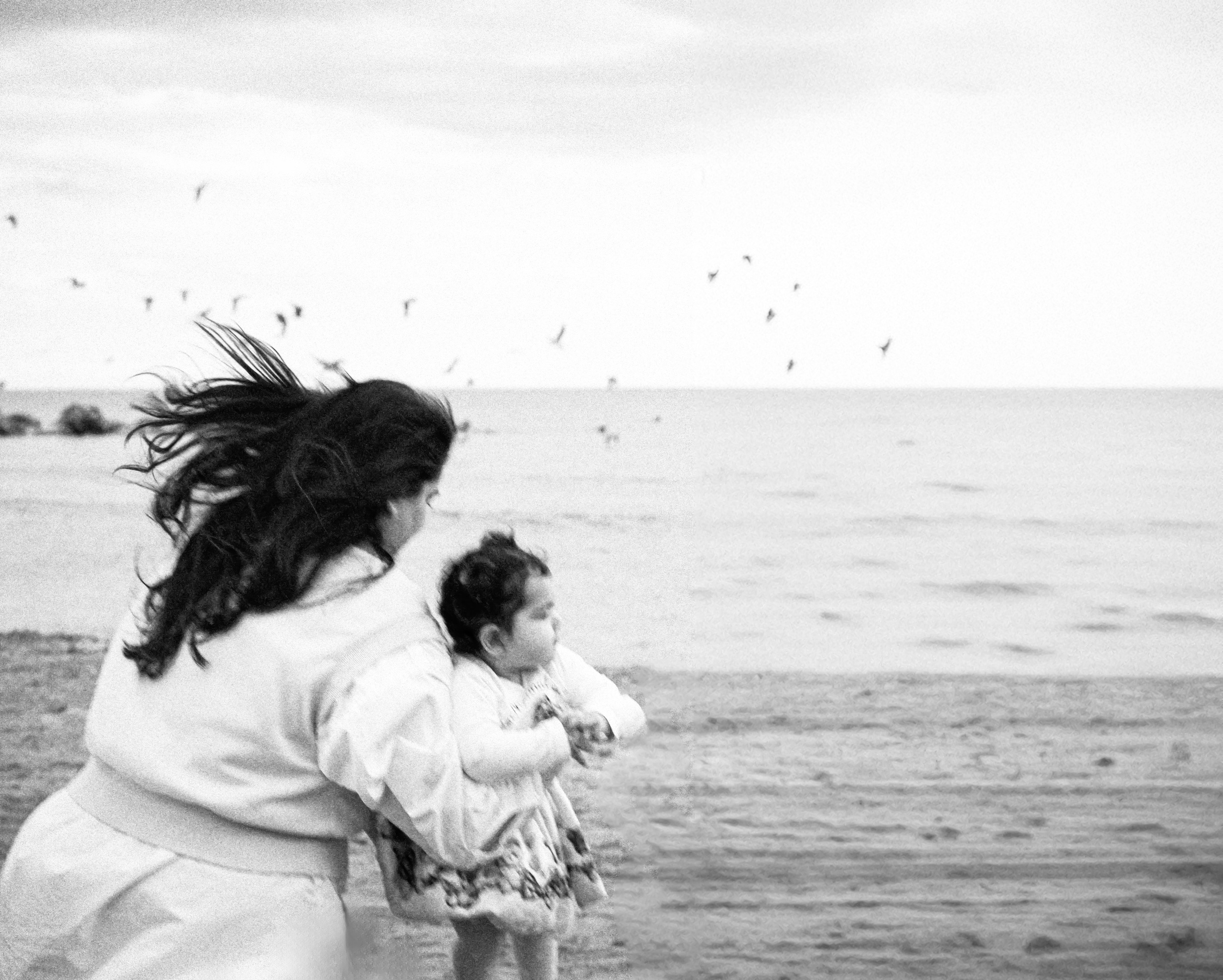 Retrato artístico en blanco y negro de madre e hija caminando por una playa ventosa con aves en vuelo y olas suaves — un momento natural y atemporal capturado en la costa de Alicante, España.