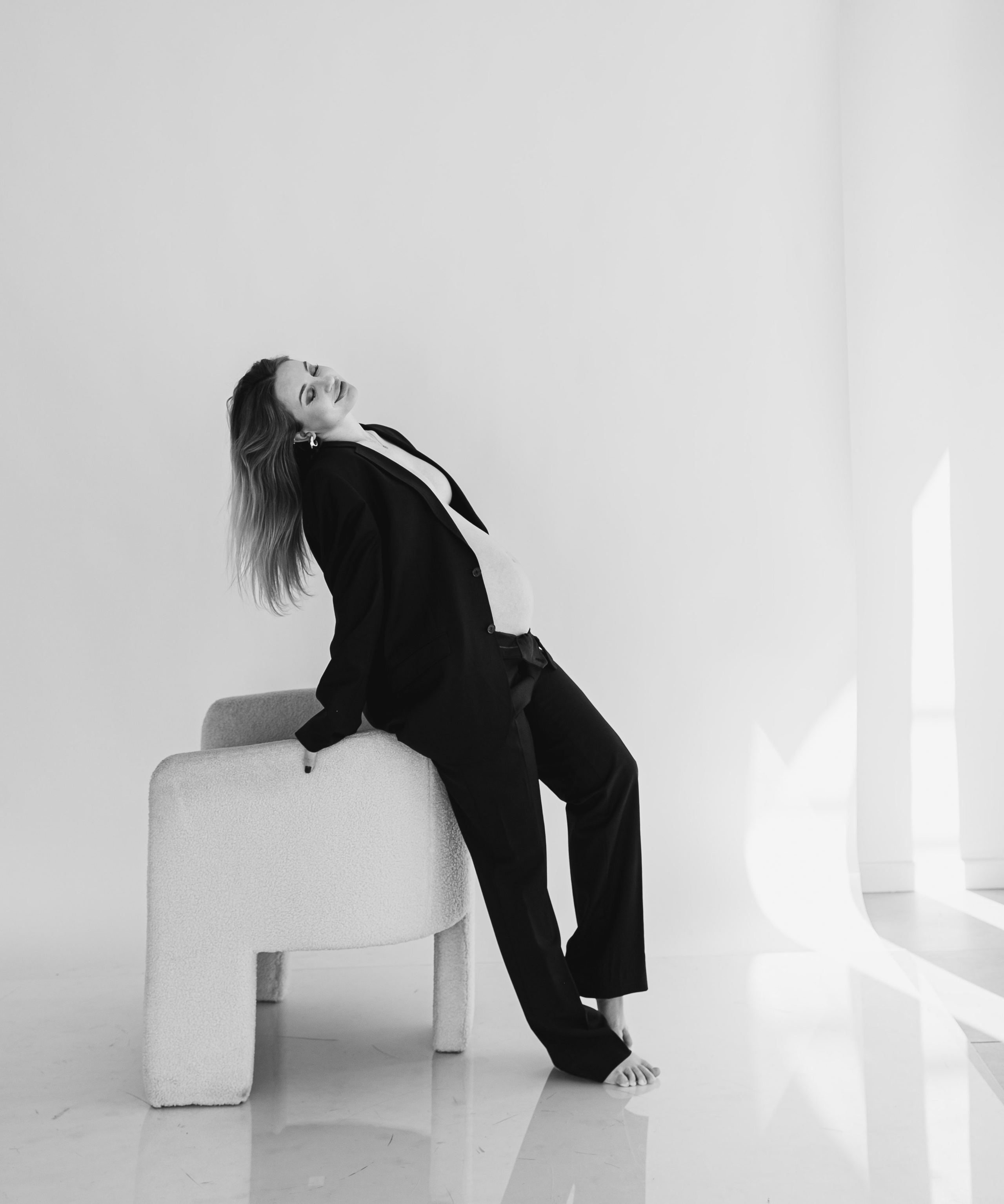 Retrato de embarazo con estilo en blanco y negro — mujer embarazada con un traje oversize, recostada con confianza sobre una silla moderna en un estudio minimalista lleno de luz natural en Valencia, España.