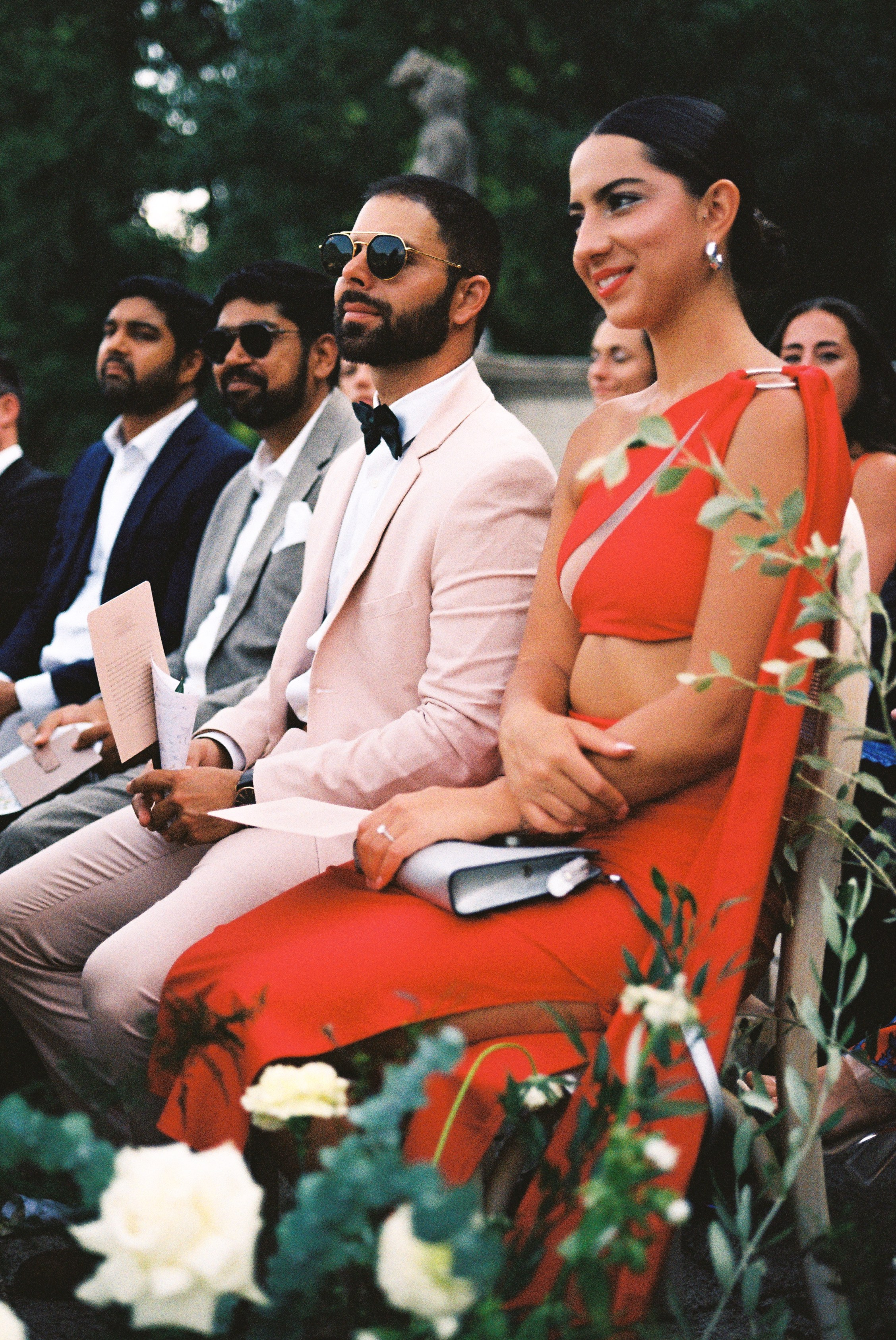 Wedding guests in stylish attire sit attentively during the ceremony, surrounded by floral decor.
