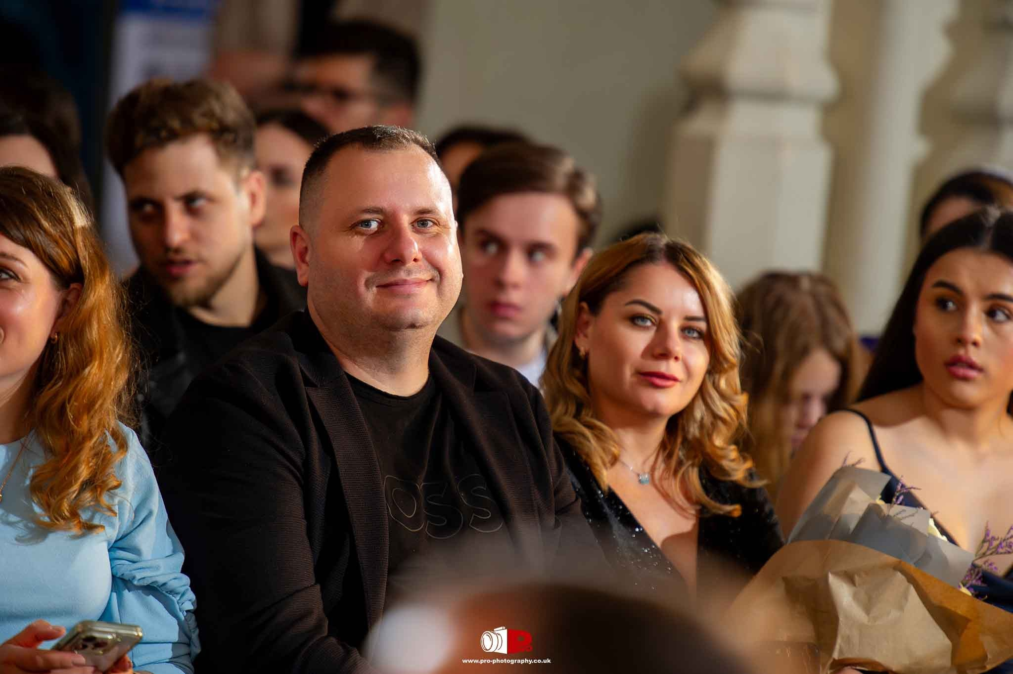 A smiling man and woman are seated among an attentive crowd at a high-profile gala event.
