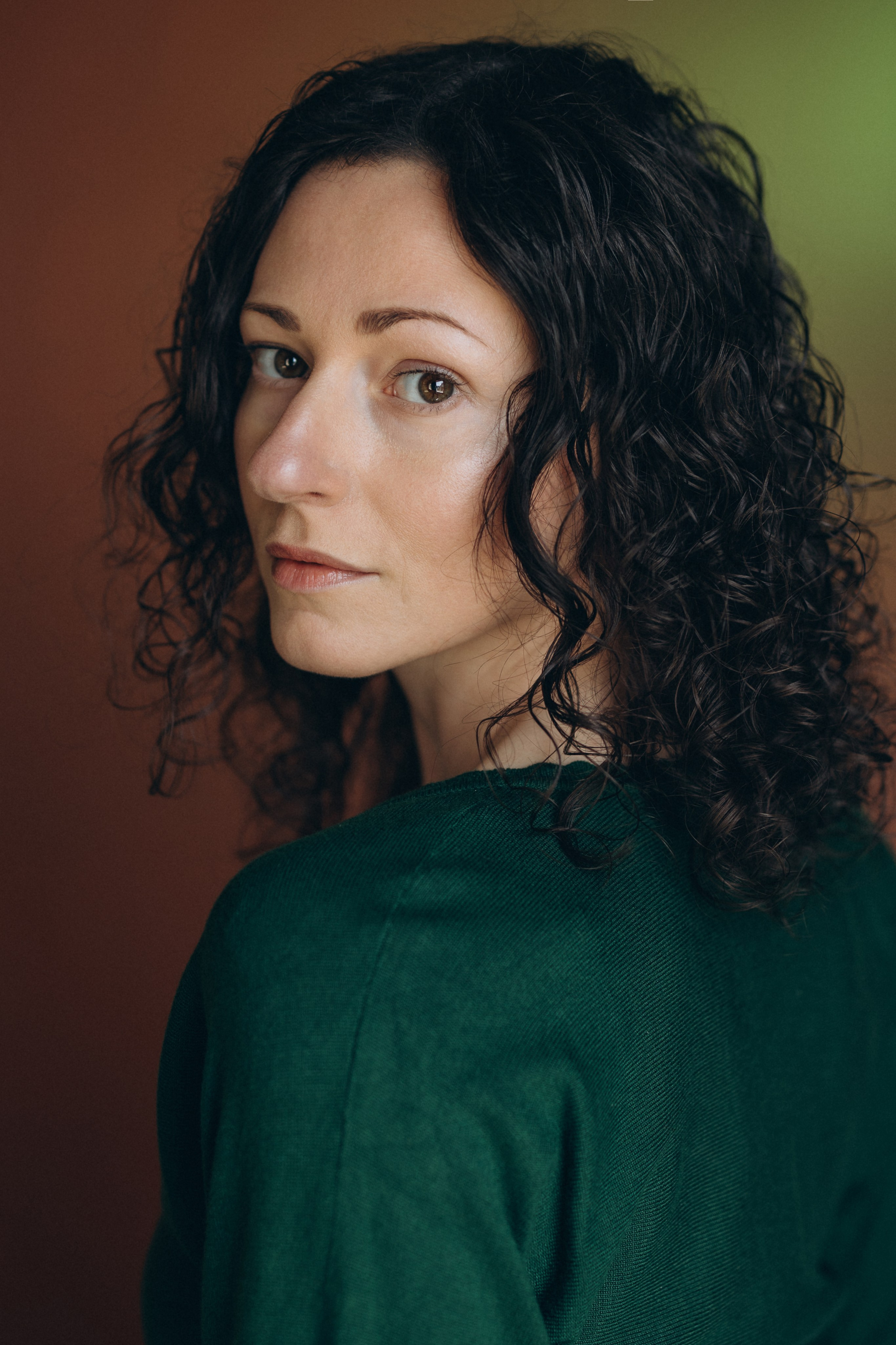Actor headshot of a woman with dark curly hair in a green top, looking to the side with a soft expression