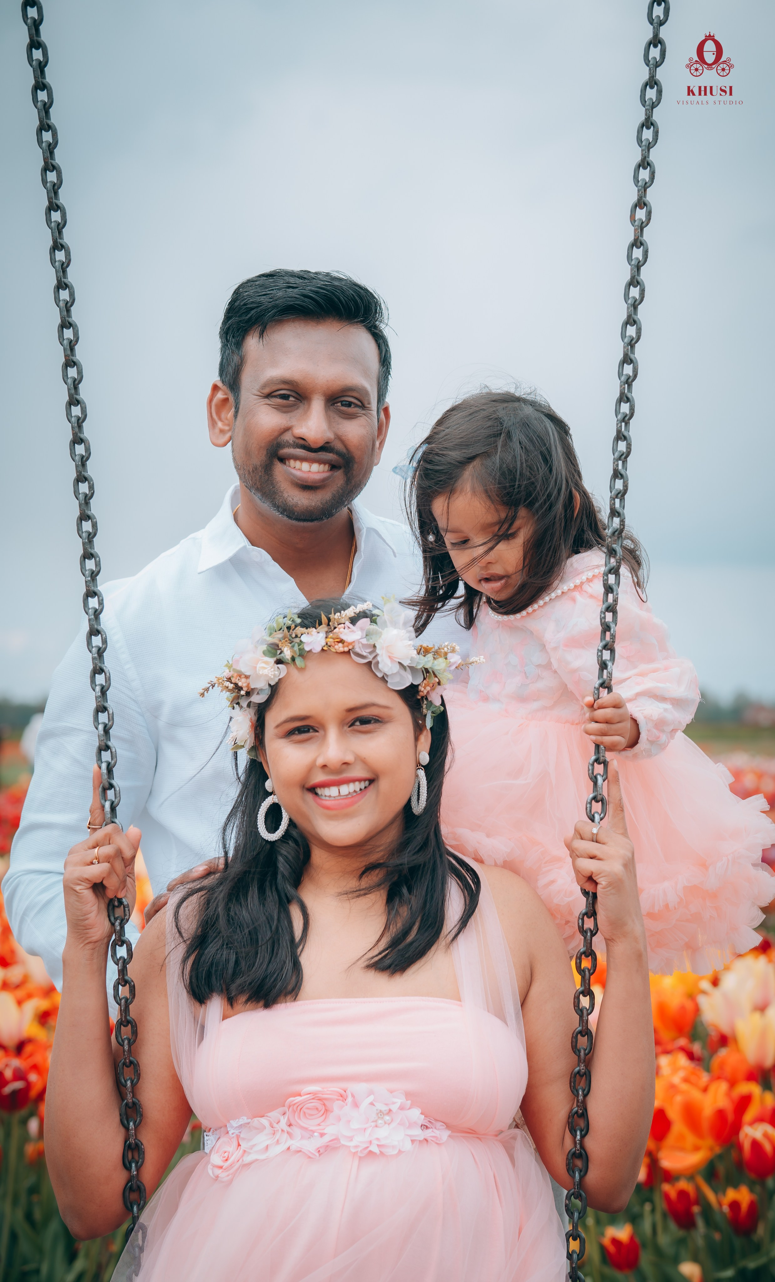 A pregnant woman sitting on a swing and her husband is holding their daughter from behind in a tulip field in netherlands
