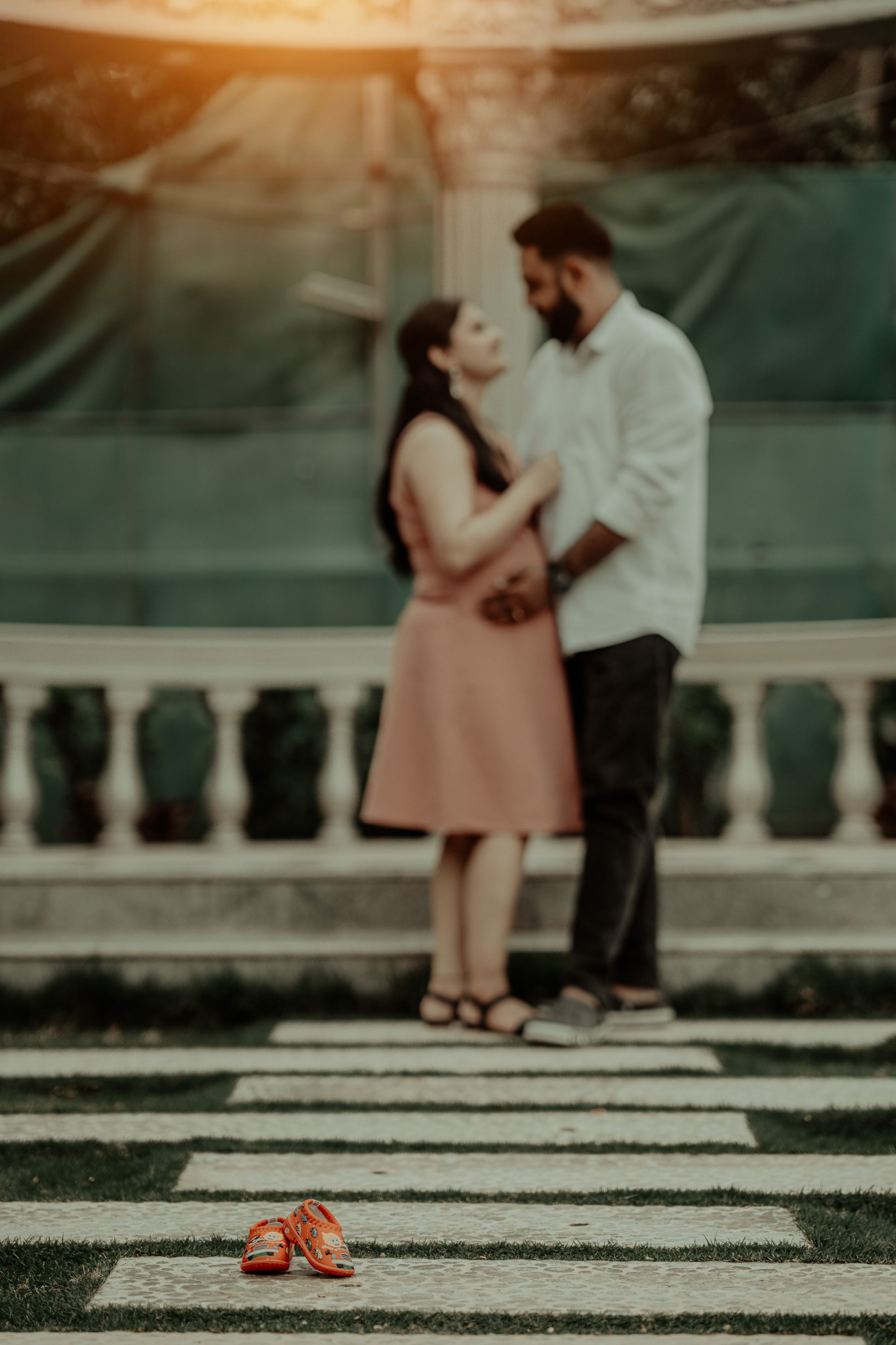 Artistic maternity photoshoot in Bengaluru featuring tiny red baby shoes in the foreground with a blurred couple standing by a white stone railing.