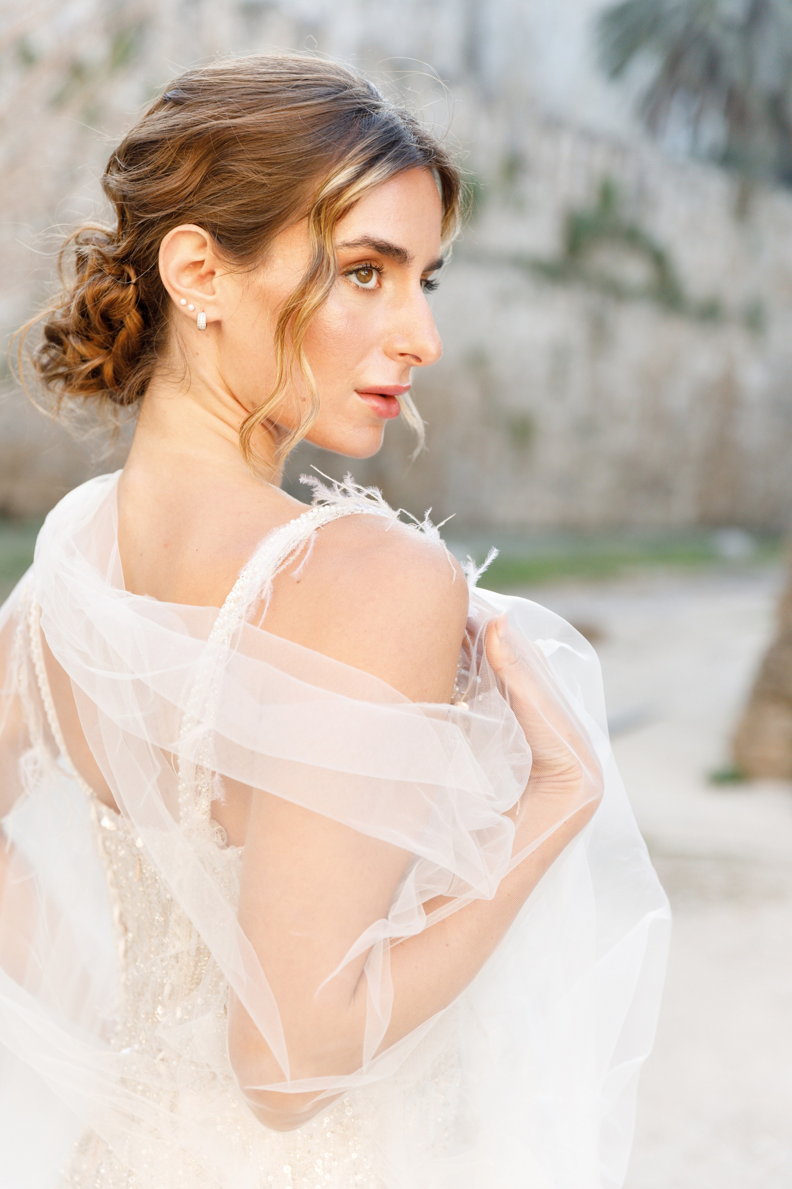 A radiant bride stands in the narrow, sunlit streets of Rhodes' Old Town, her intricate lace wedding dress contrasting beautifully with the weathered stone walls behind her. The editorial-style portrait highlights her serene expression and the timeless elegance of the historic setting, with soft natural light accentuating her features.
