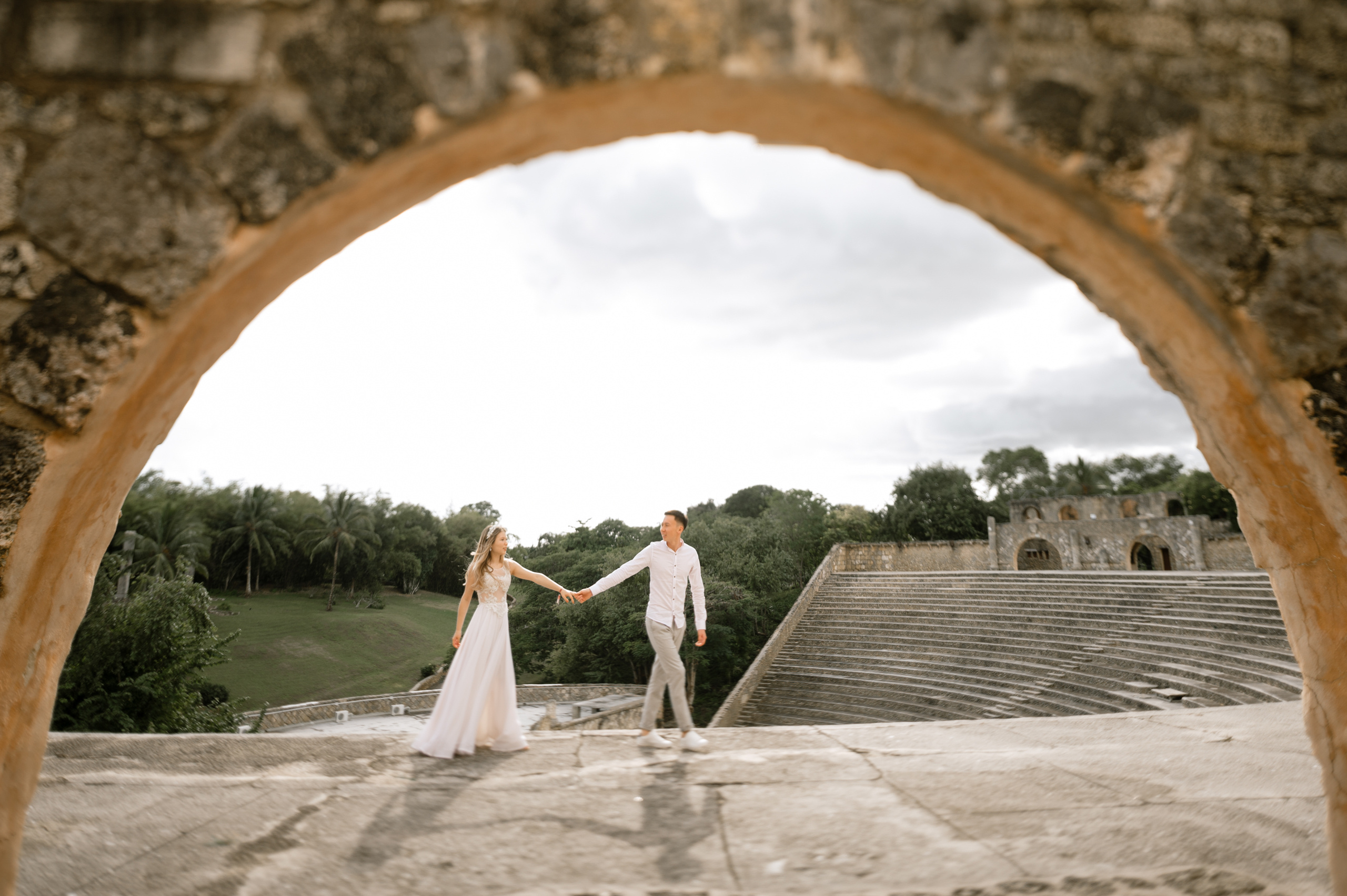 Через каменную арку видны жених и невеста на фоне арочной каменной лестницы и зеленой горы.