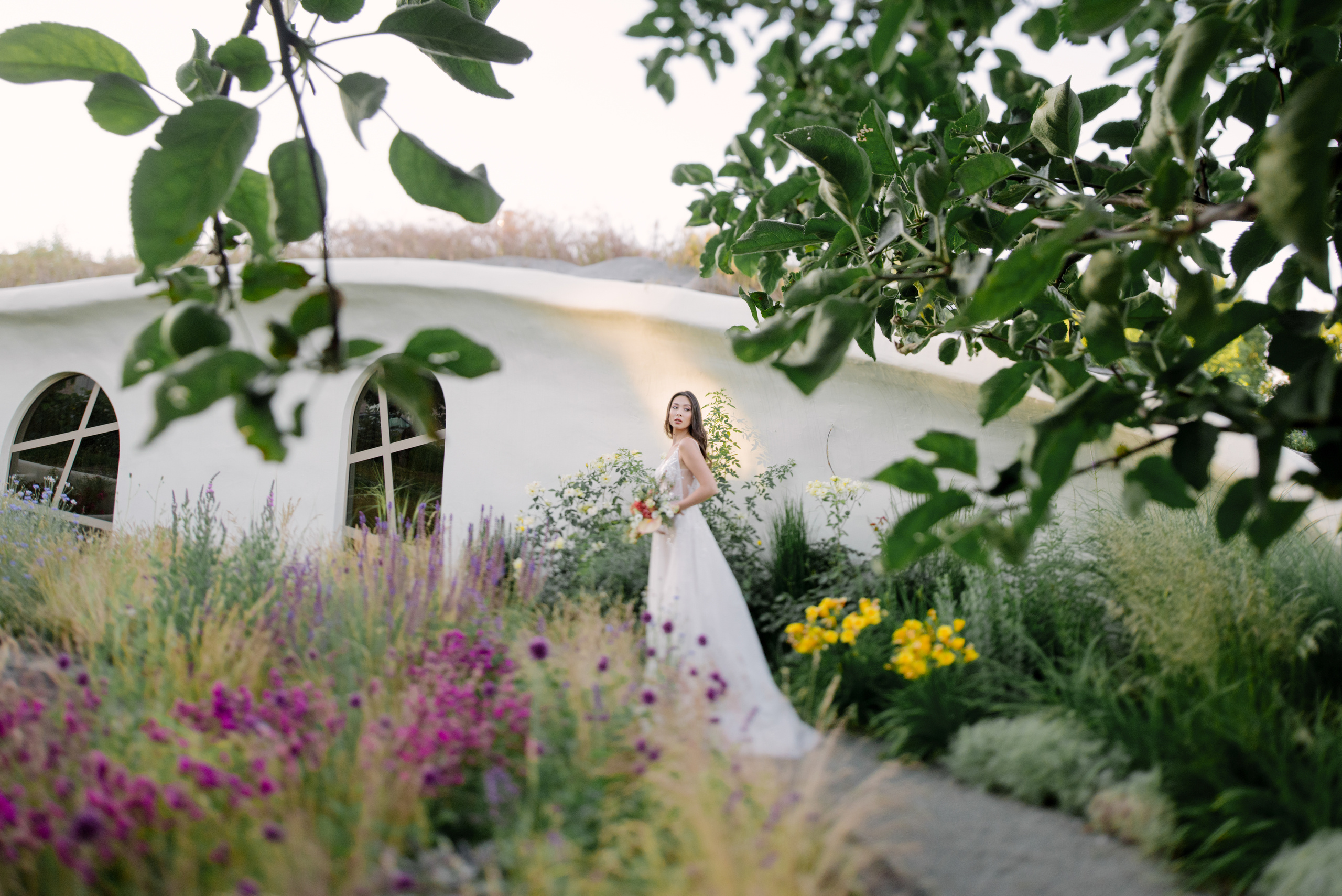 A bride in a white wedding dress with a bouquet of wildflowers stands on the path against the background of a fairy-tale house.