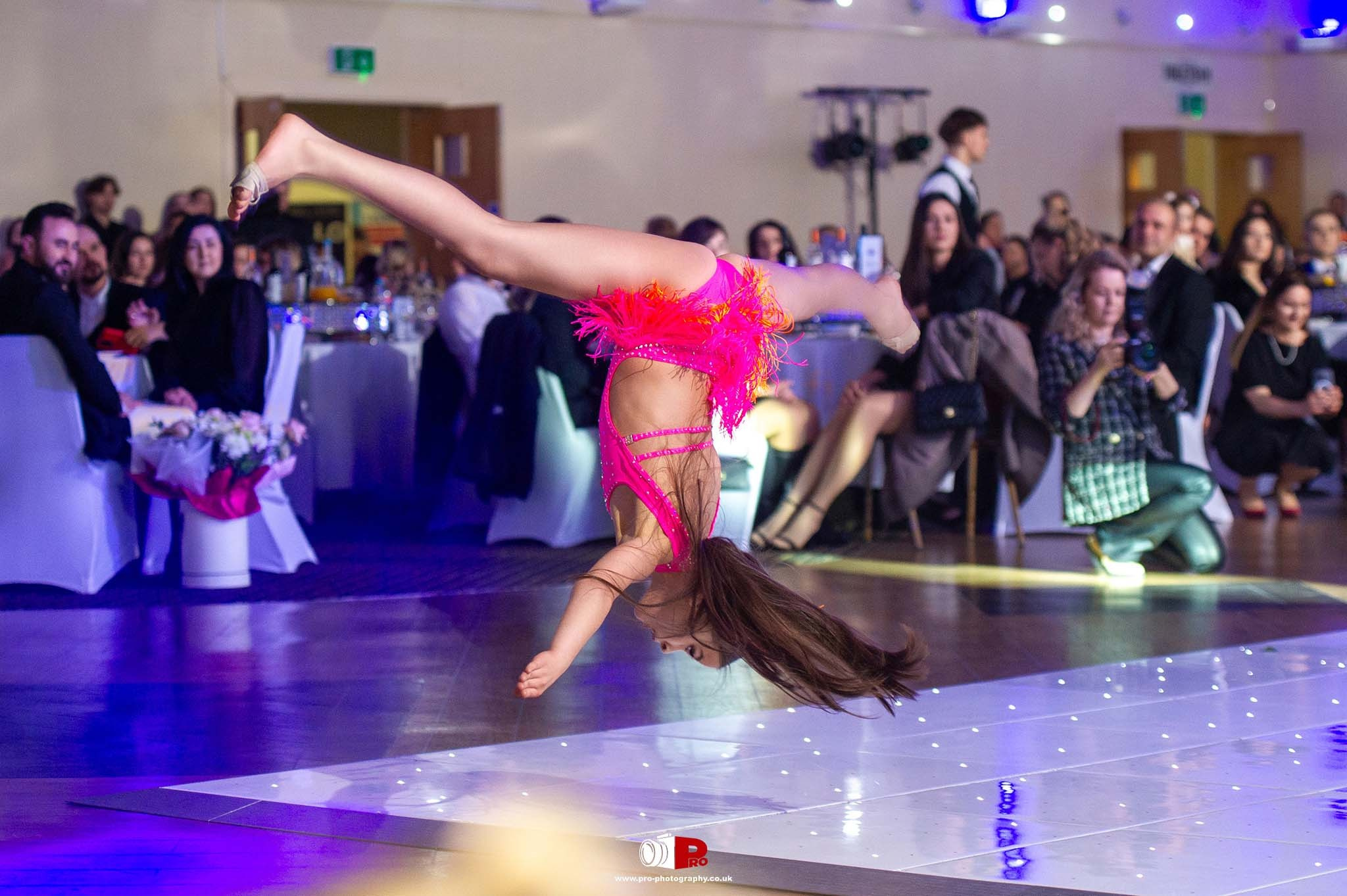 A young dancer in a vibrant pink outfit performs a stunning mid-air acrobatic move during a gala event.