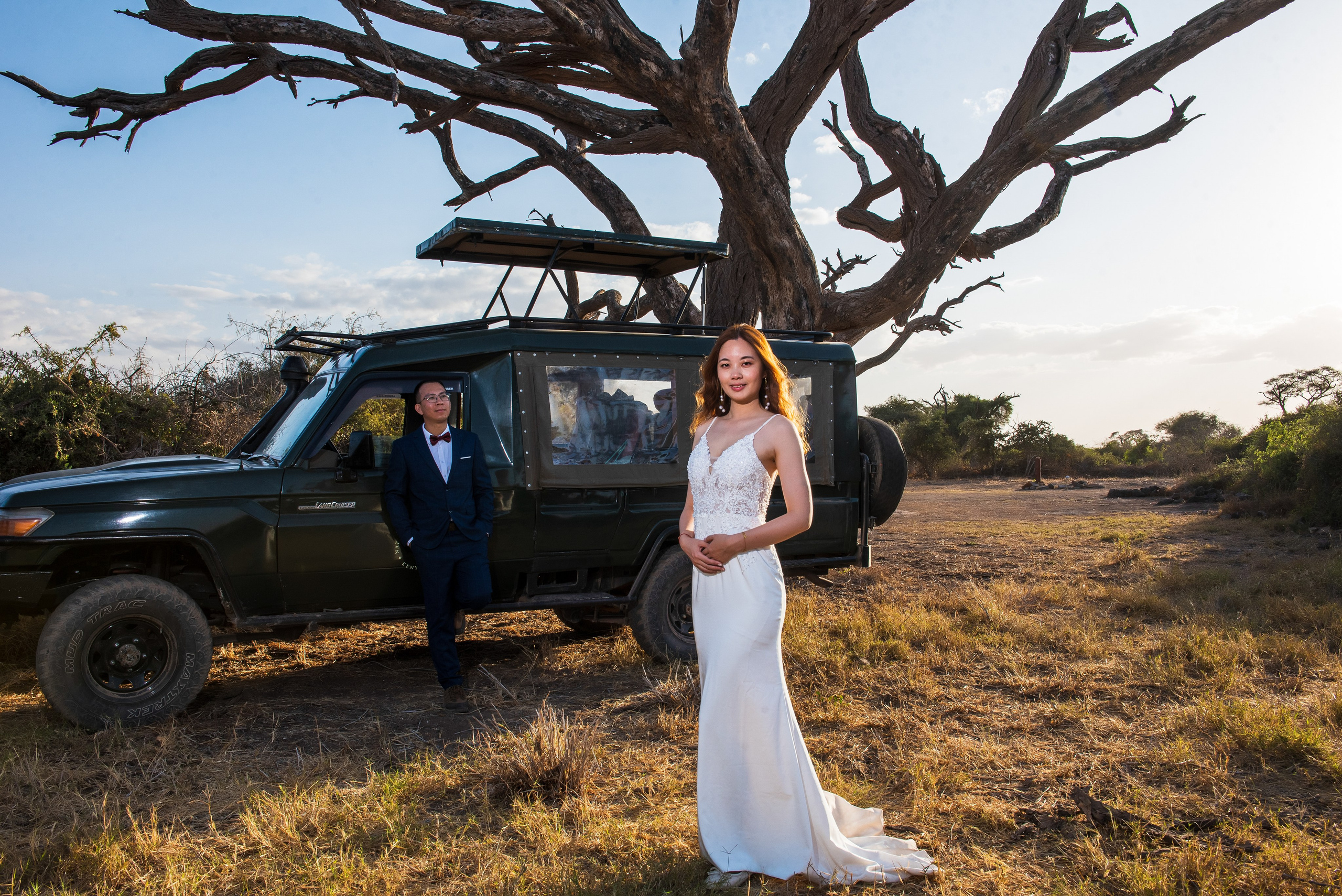 A wide shot of a Chinese bride and groom, next to a tours & travel landcruiser, in Amboseli, during their wedding photo session.