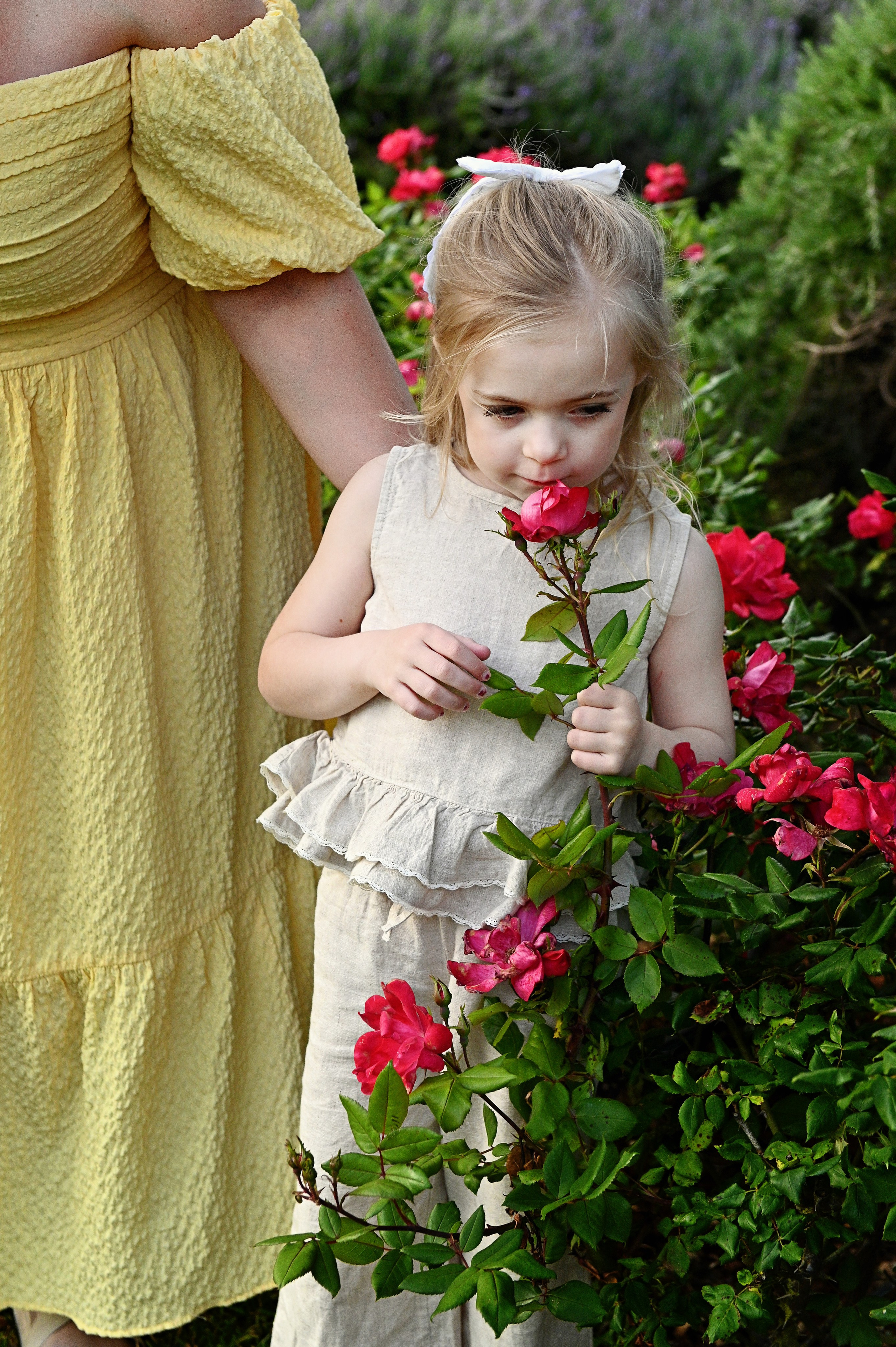 Servizio fotografico per bambini in Puglia: una bambina in un completo di lino crema tiene in mano e annusa una rosa rossa accanto a una donna in abito giallo.