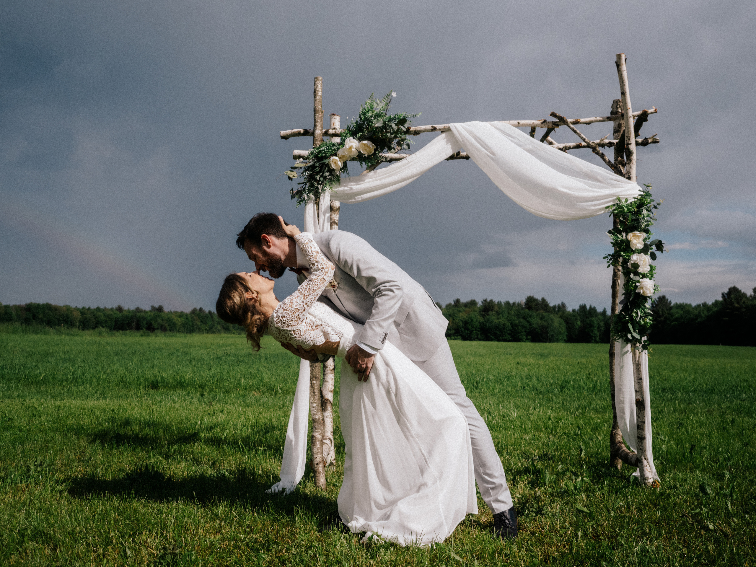 Couple in wedding attire dip kissing in front of wide field and a rainbow in Saint Thomas, Quebec.