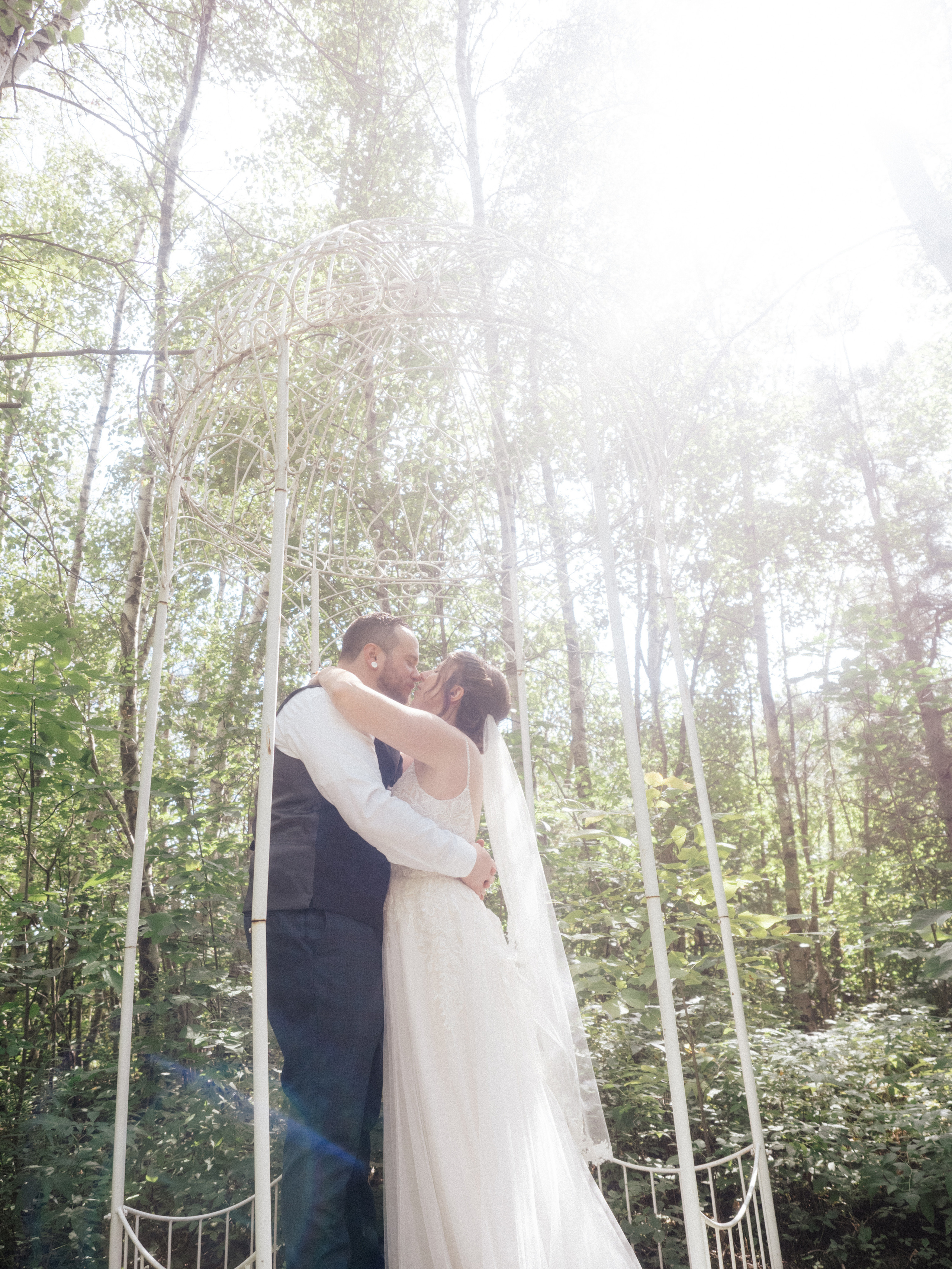Couple in wedding attire kissing in forest with light coming through trees on Domaine St-Simon site in Saint-Simon-de-Bagot, Quebec.