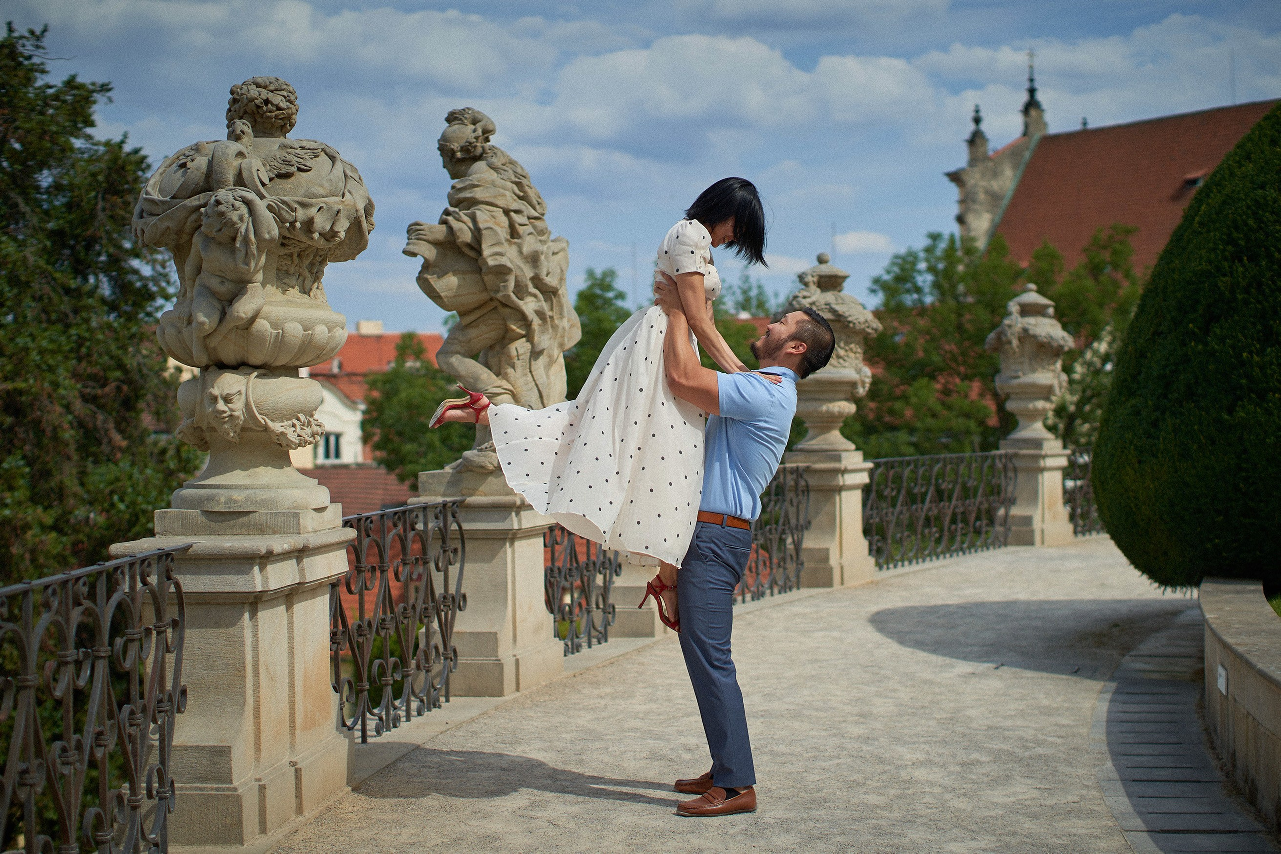 Man lifting his fiancée aloft surrounded by classical statues in the middle terrace of Vrtba Garden.