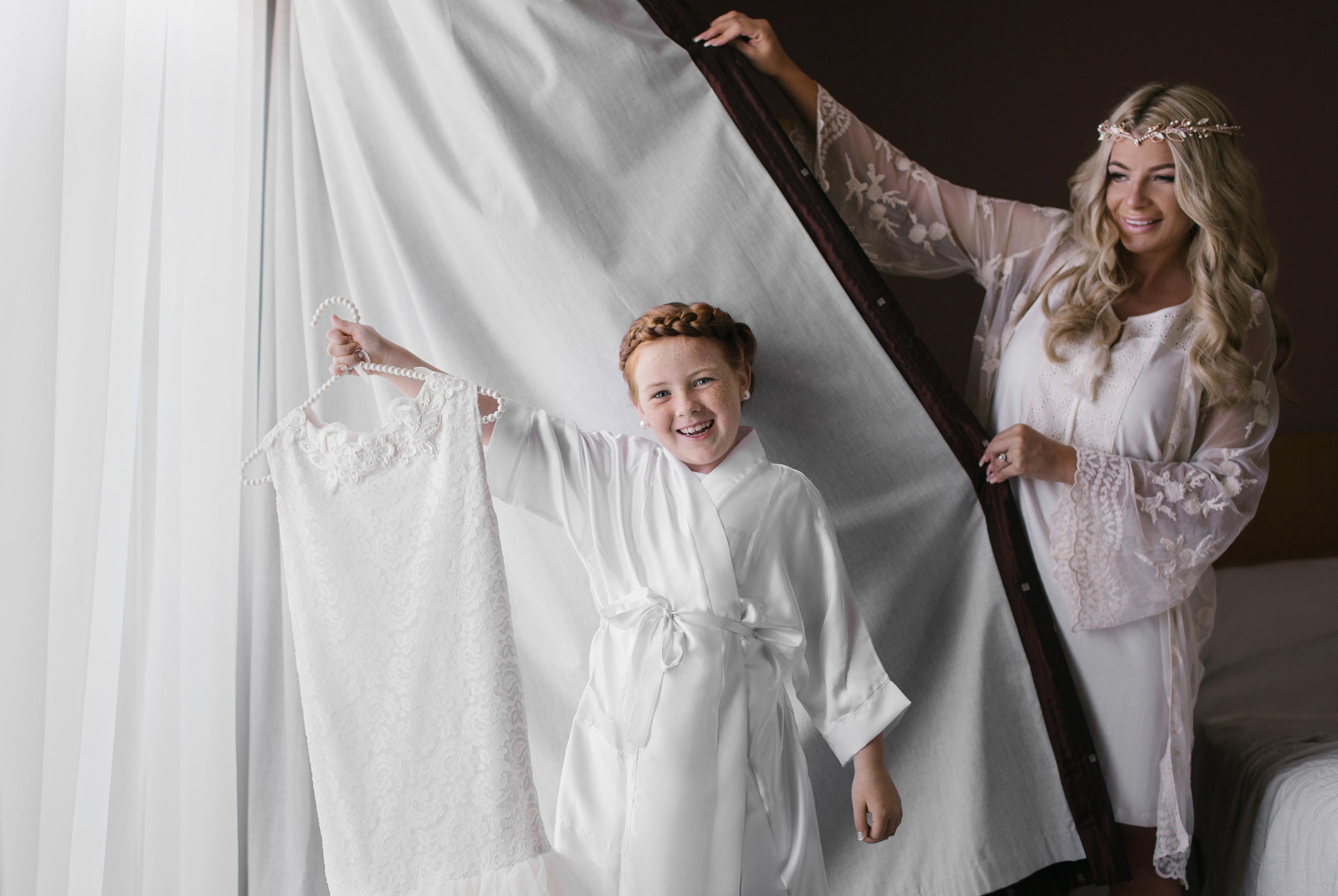 red hair girl is holding her white dress and beautiful bride is admiring her