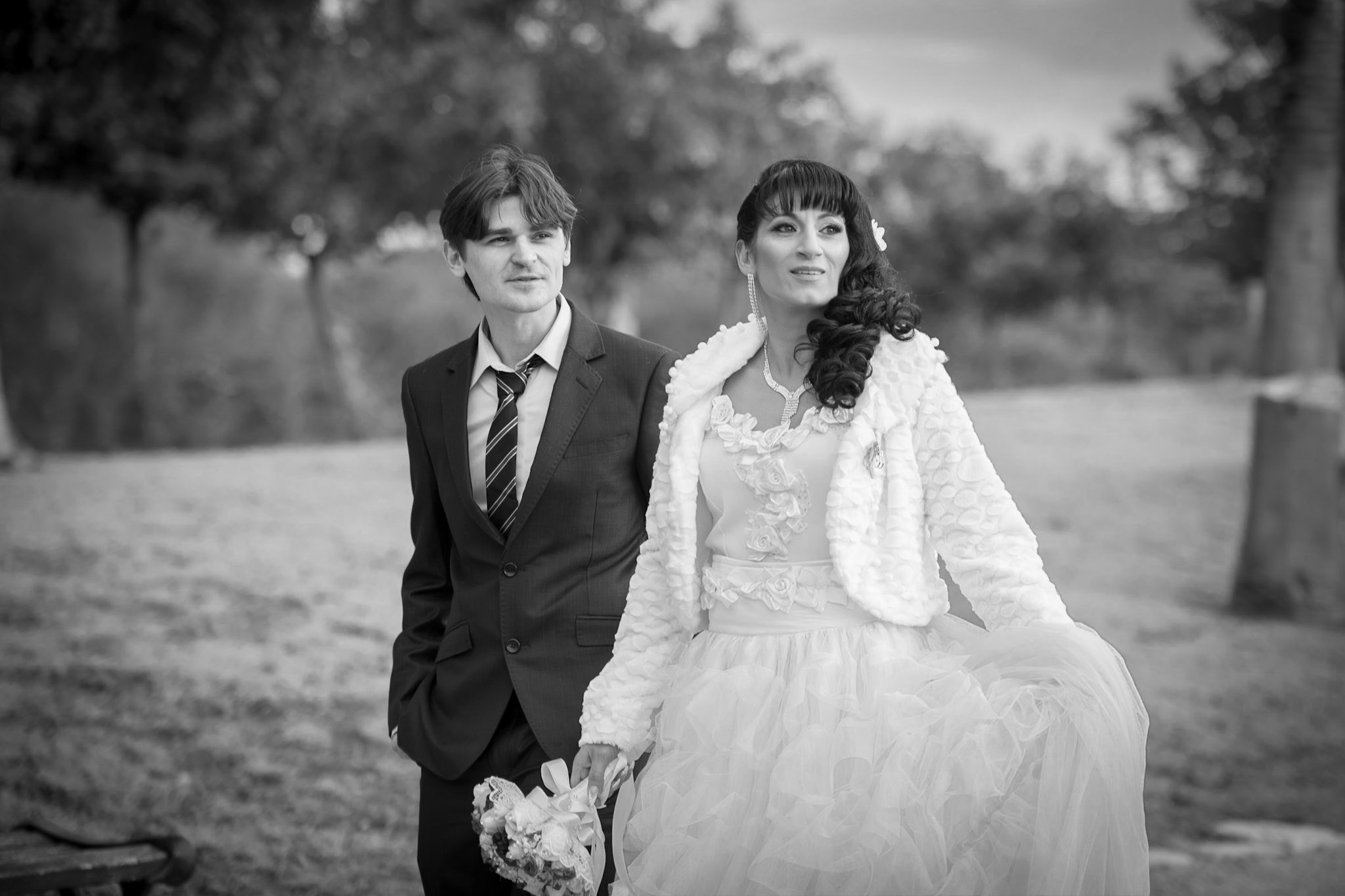 A black-and-white image capturing a wedding stroll. The bride, in a voluminous white dress and jacket with flowers in her hair, and the groom, in a dark suit with a tie, walk side by side outdoors, in a park, under overcast weather. The background is blurred, focusing attention on the couple at the center of the frame. The photo style is black-and-white, creating a retro and elegant atmosphere.