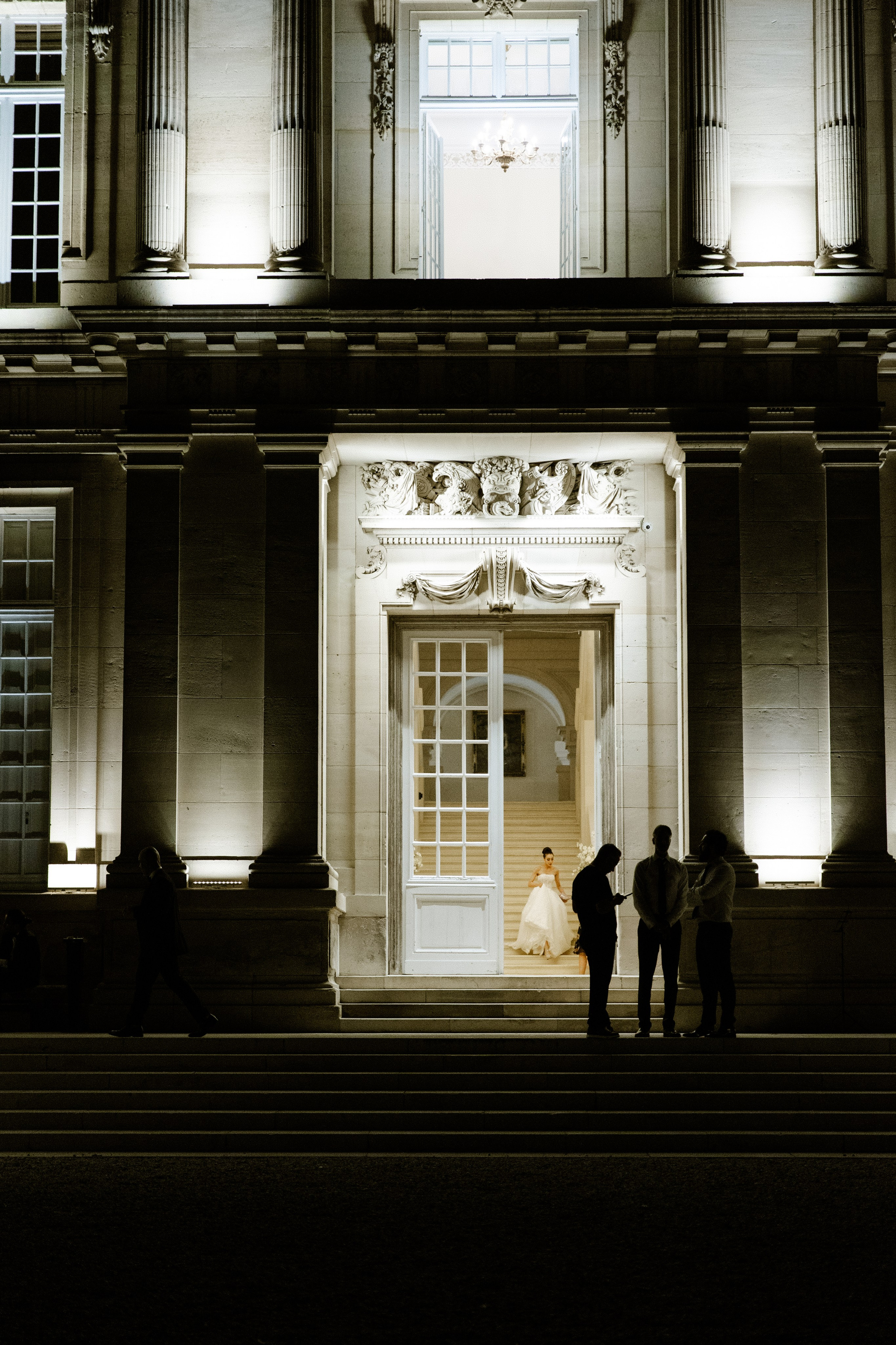 Elegant bride standing at the illuminated entrance of a French château at night, captured in a cinematic wedding style