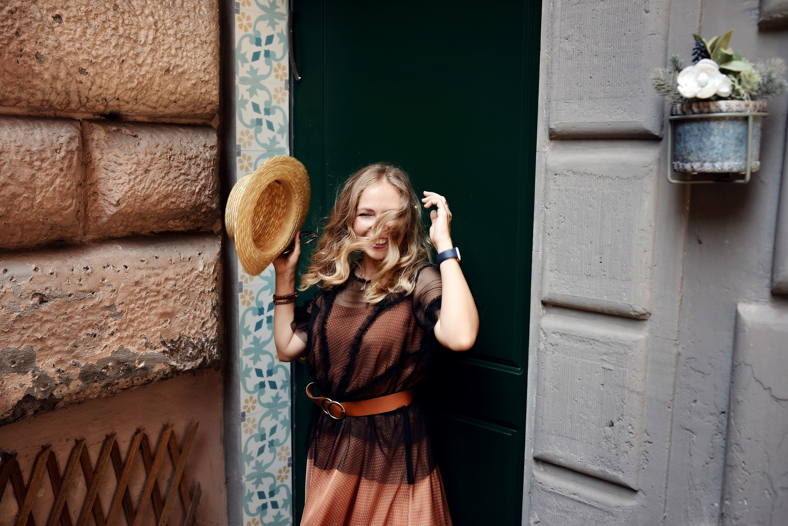 A cheerful young woman in a stylish dress plays with her straw hat as the wind blows through her hair in front of a dark green door.