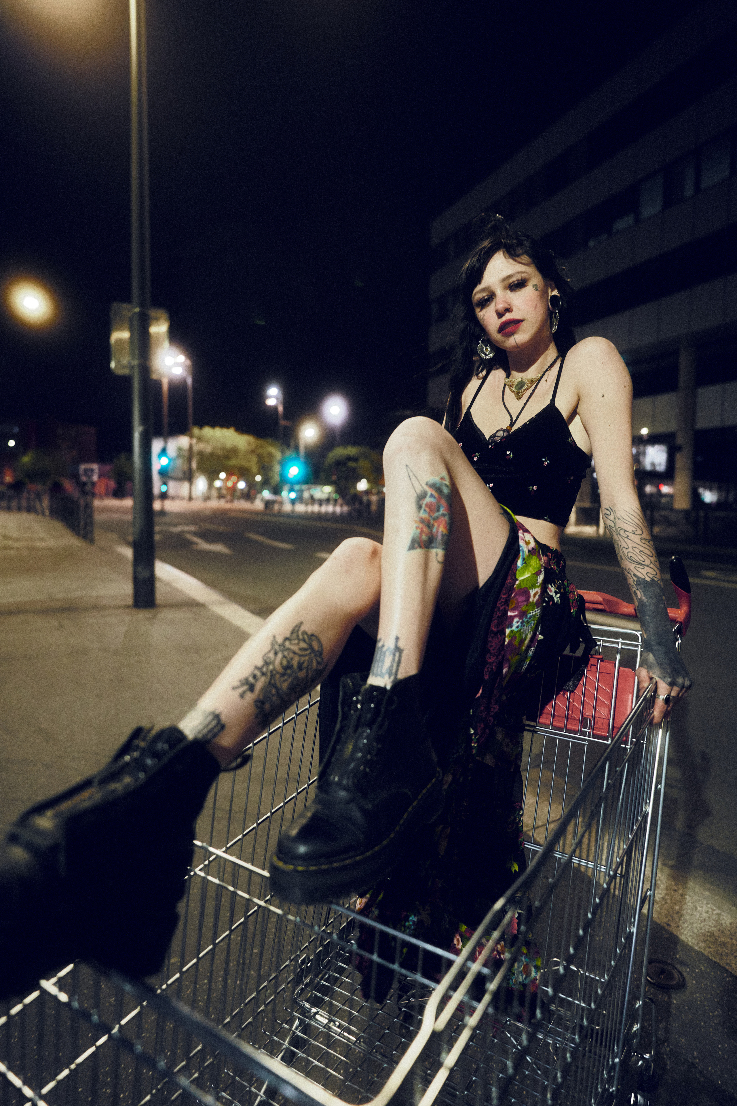  A woman sitting in a shopping cart on a city street at night, wearing edgy clothing and exuding a rebellious vibe.