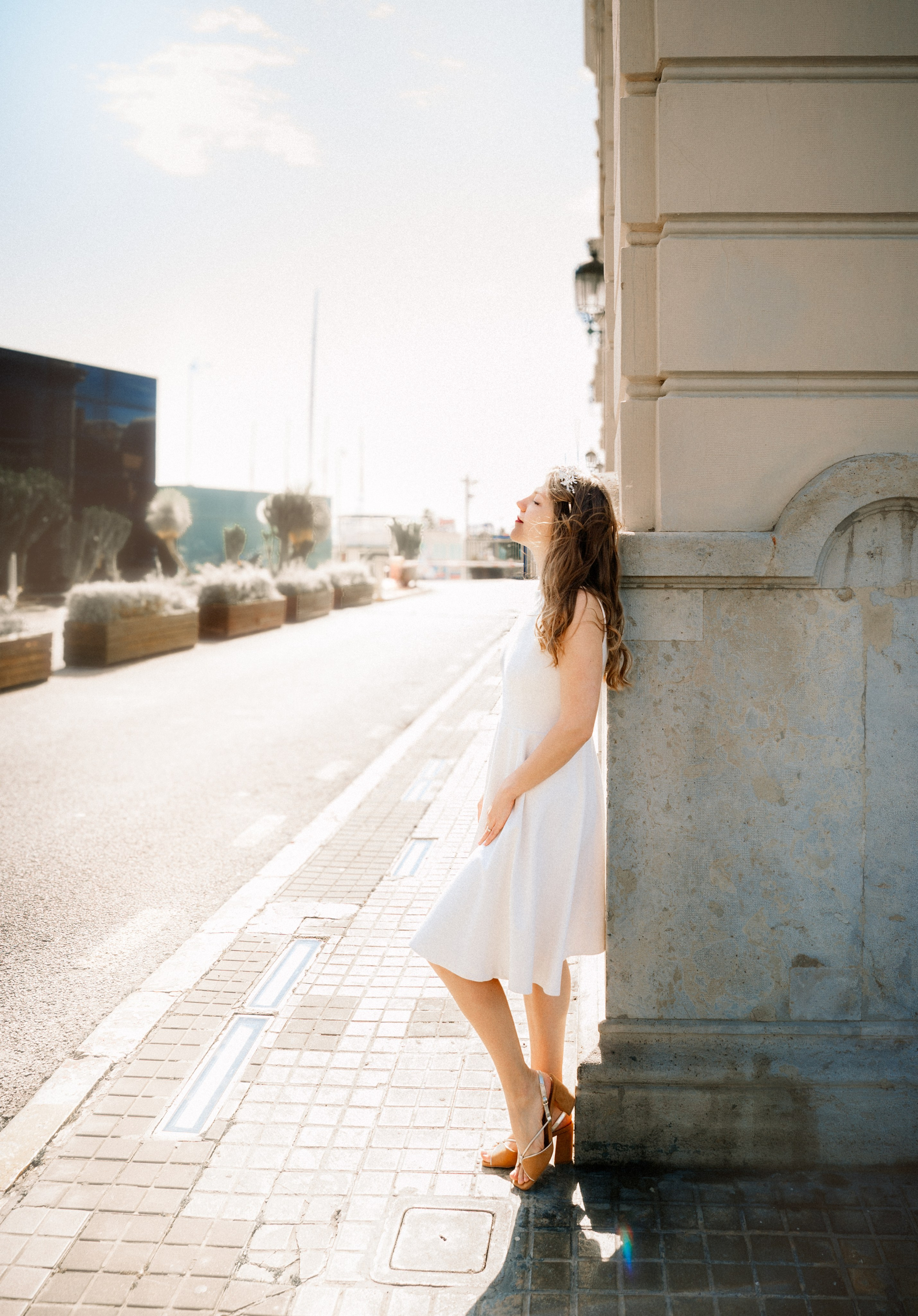 Мечтательный уличный портрет женщины в белом платье, прислонившейся к освещённой солнцем каменной стене на тихой улочке в Кальпе, Испания — идеально подходит для романтических и светлых портретных фотосессий в прибрежных испанских городках.