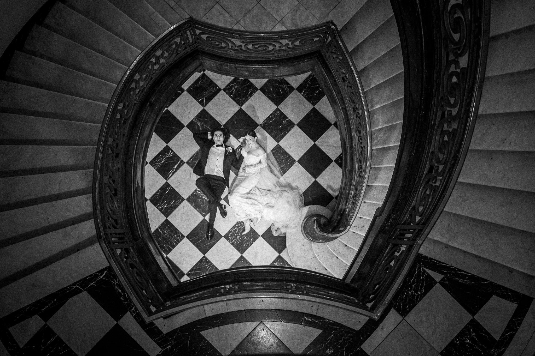 Elegantna crno-bela fotografija venčanja: mladenci leže na spiralnom stepeništu sa dezenom šahovske table na podu.