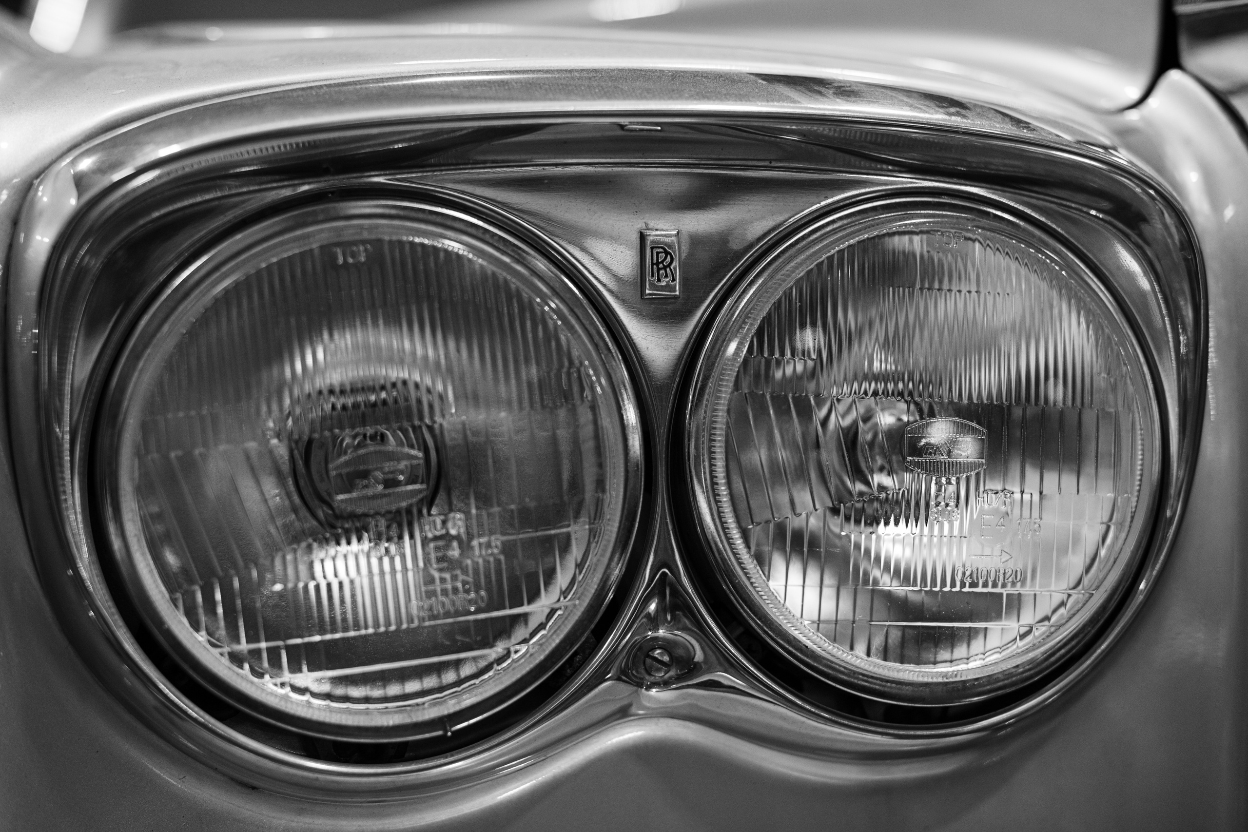 Detailed black-and-white close-up of a classic Rolls-Royce dual headlight with the signature Rolls-Royce emblem centered between the lights. This photo, captured at The Garage by Classic Motorworks in Singapore, emphasizes luxury car craftsmanship and precision design, showcasing MirrorKlezz Photography’s skill in capturing the fine details of vintage automobiles. https://www.classicmotorworks.com.sg/