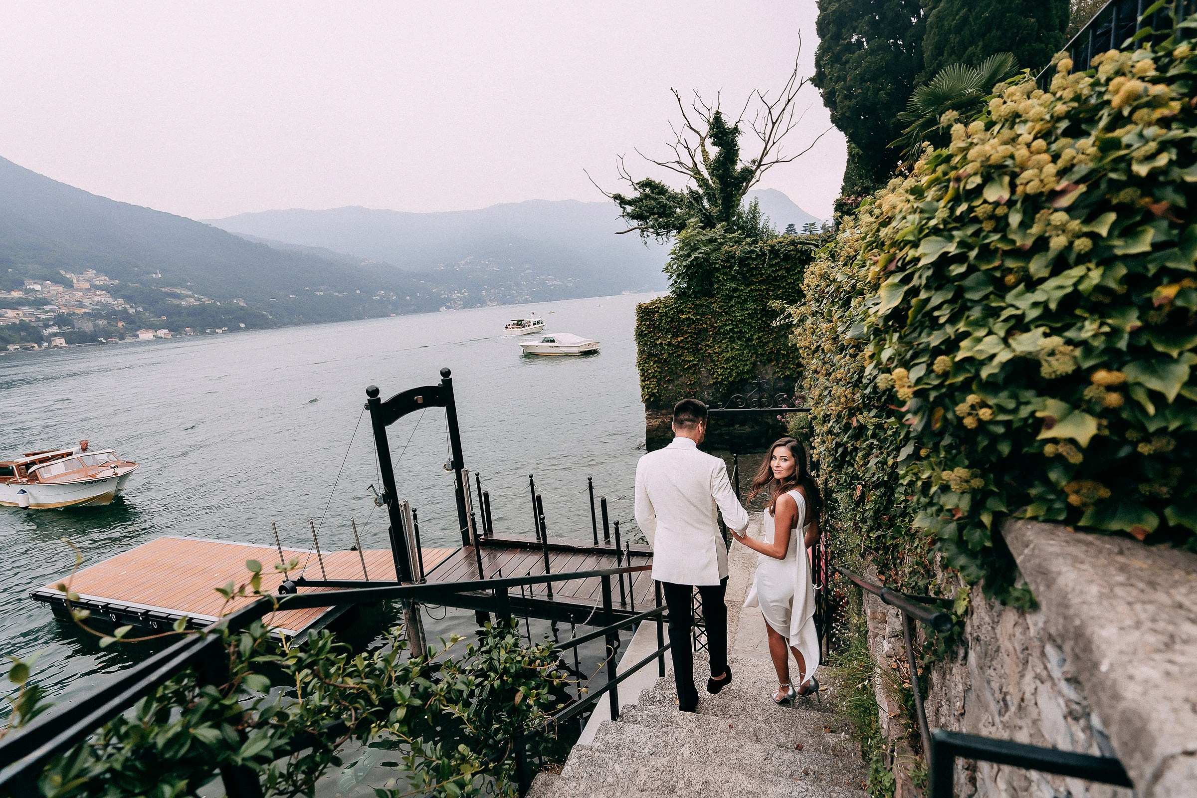 Elegant couple descending stone steps toward lakeside dock with scenic mountain views and boats, surrounded by lush greenery on a romantic getaway.