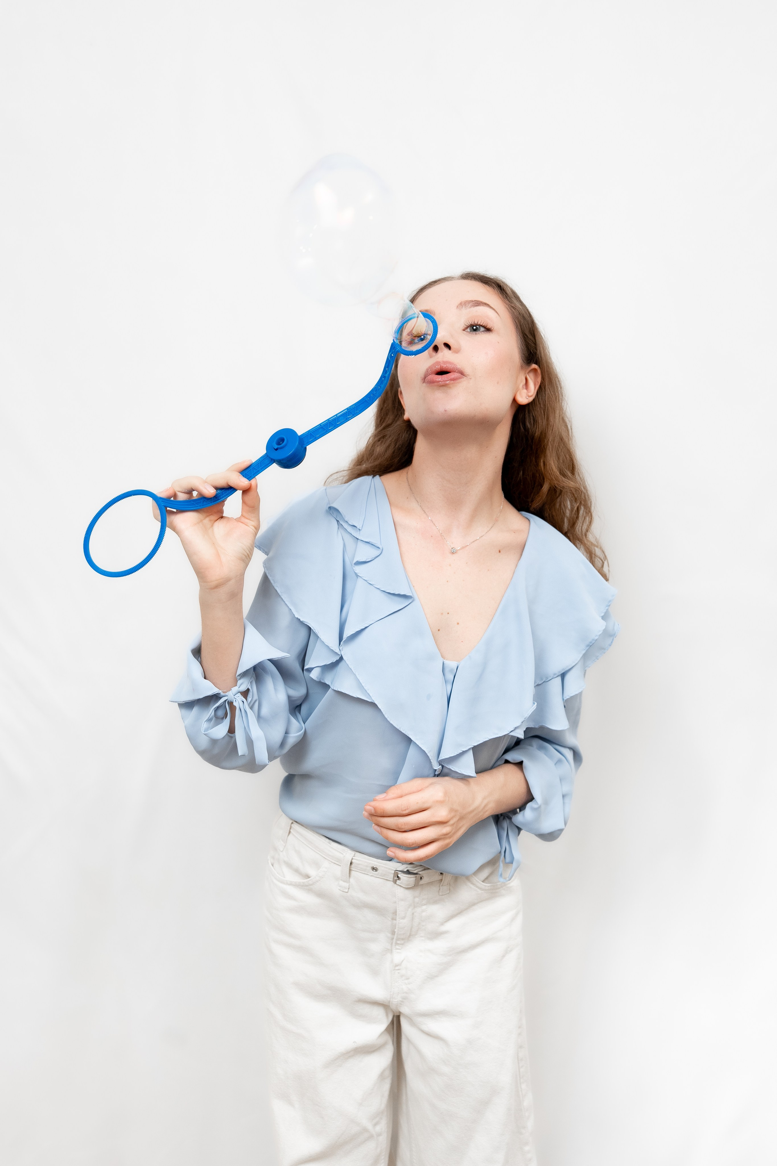 Young woman in a light blue blouse blowing a soap bubble with a large blue bubble wand on a white background.
