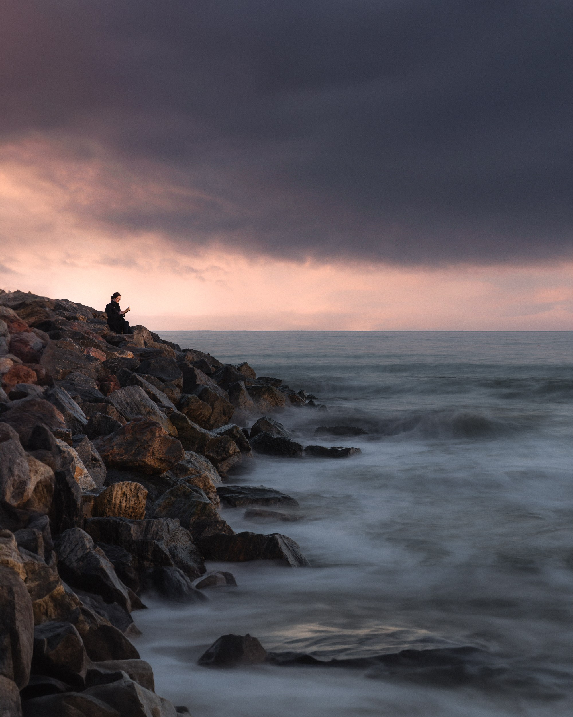 A photo of a woman reading a book on the rocks next to the sea 