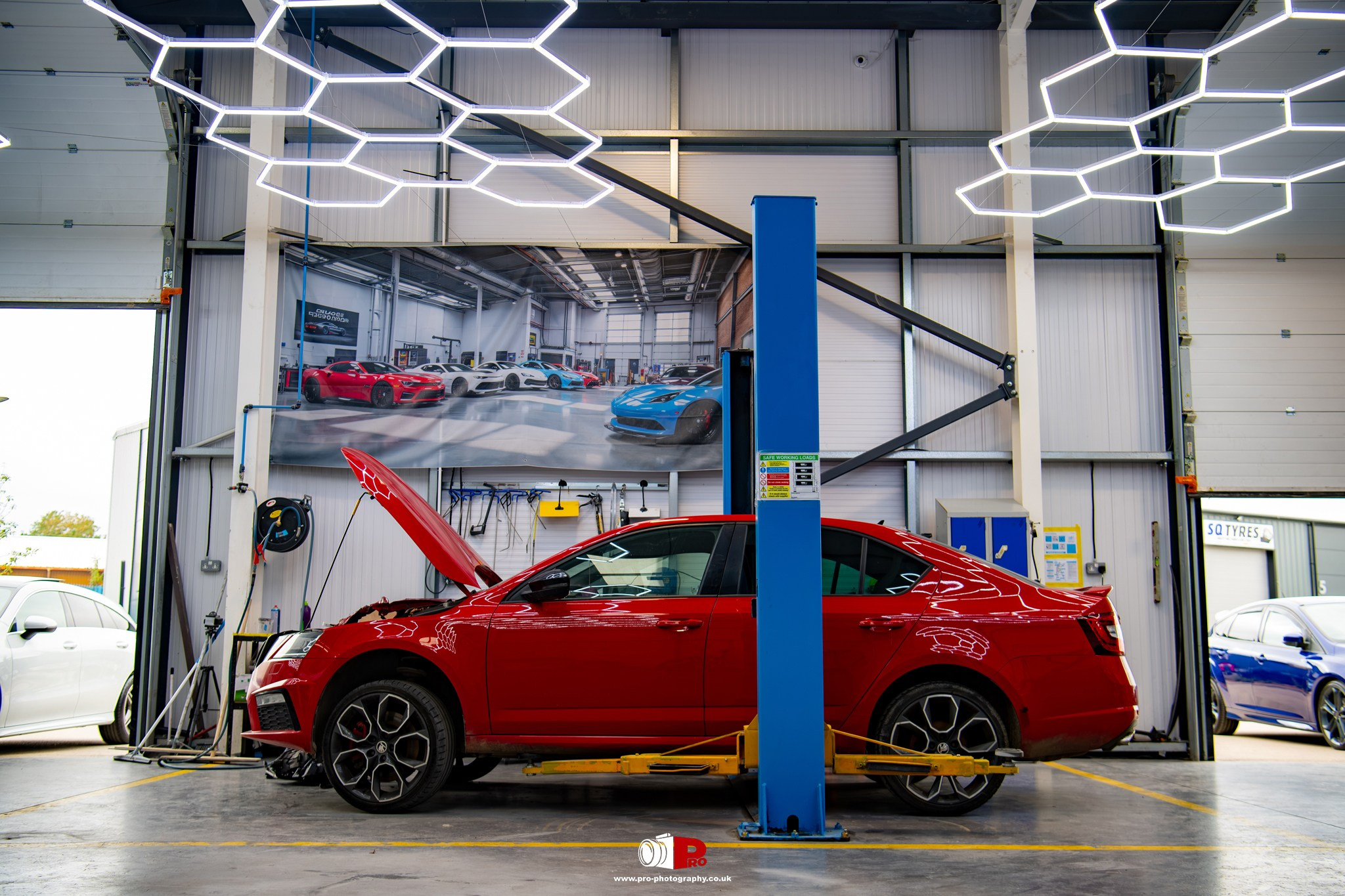 Commercial automotive image of a red car raised on a vehicle lift inside a professional body repair workshop.