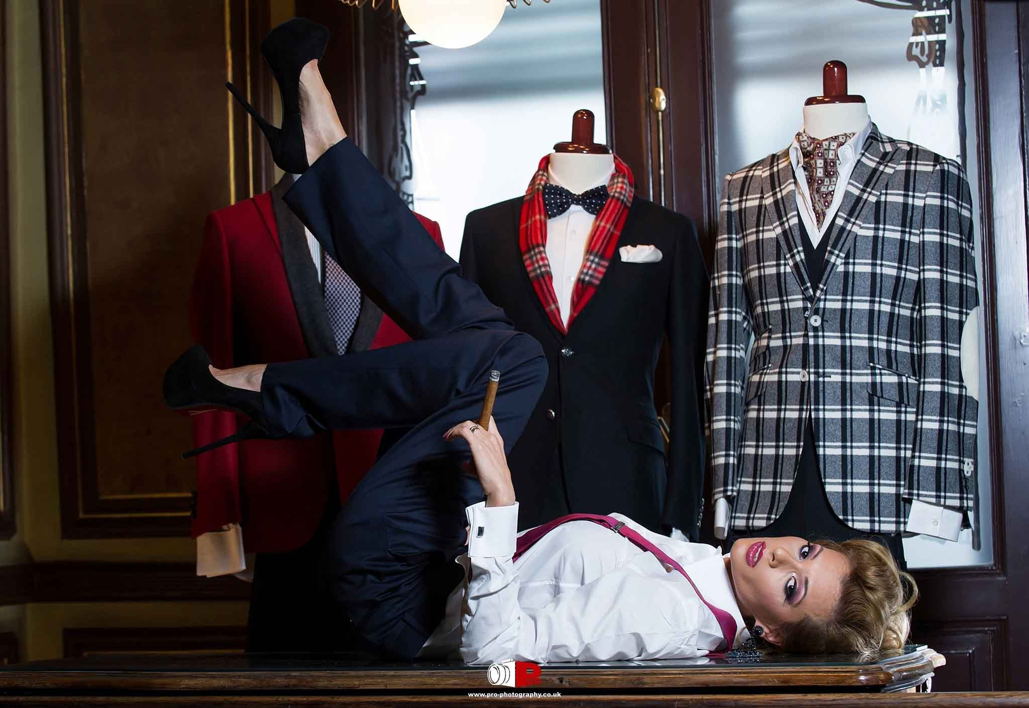 Creative fashion advertising photograph featuring a woman posing among tailored menswear displays inside a clothing boutique.