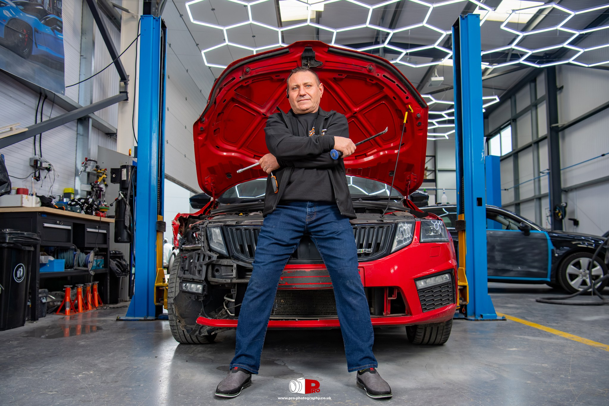 Commercial environmental portrait of an automotive repair professional standing in front of a damaged red car inside a body shop.