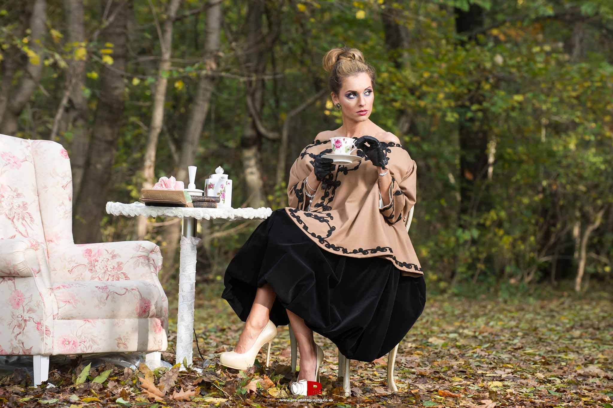 Styled outdoor fashion portrait of a woman seated with a tea set in a woodland setting.