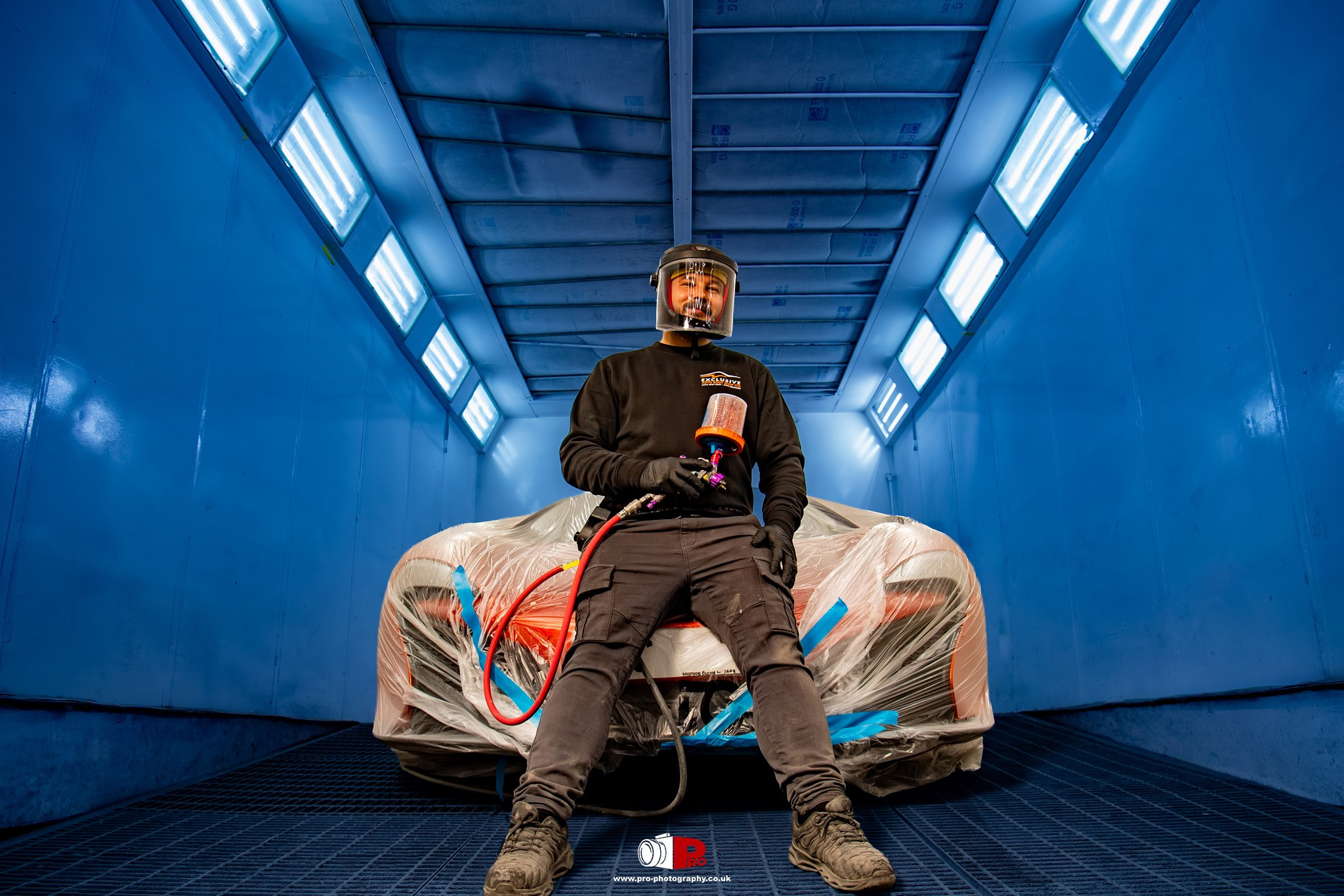 Commercial automotive image of a vehicle painter inside a professional spray booth preparing a masked car for refinishing.