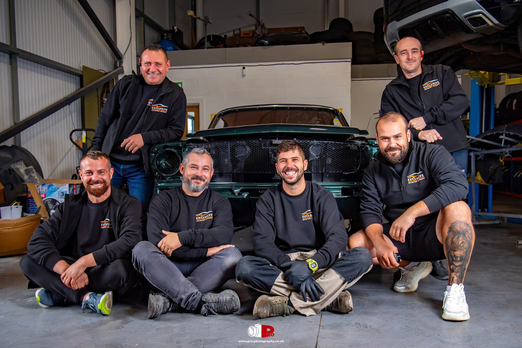 Group portrait of an automotive repair team posing together around a classic car inside a professional workshop.
