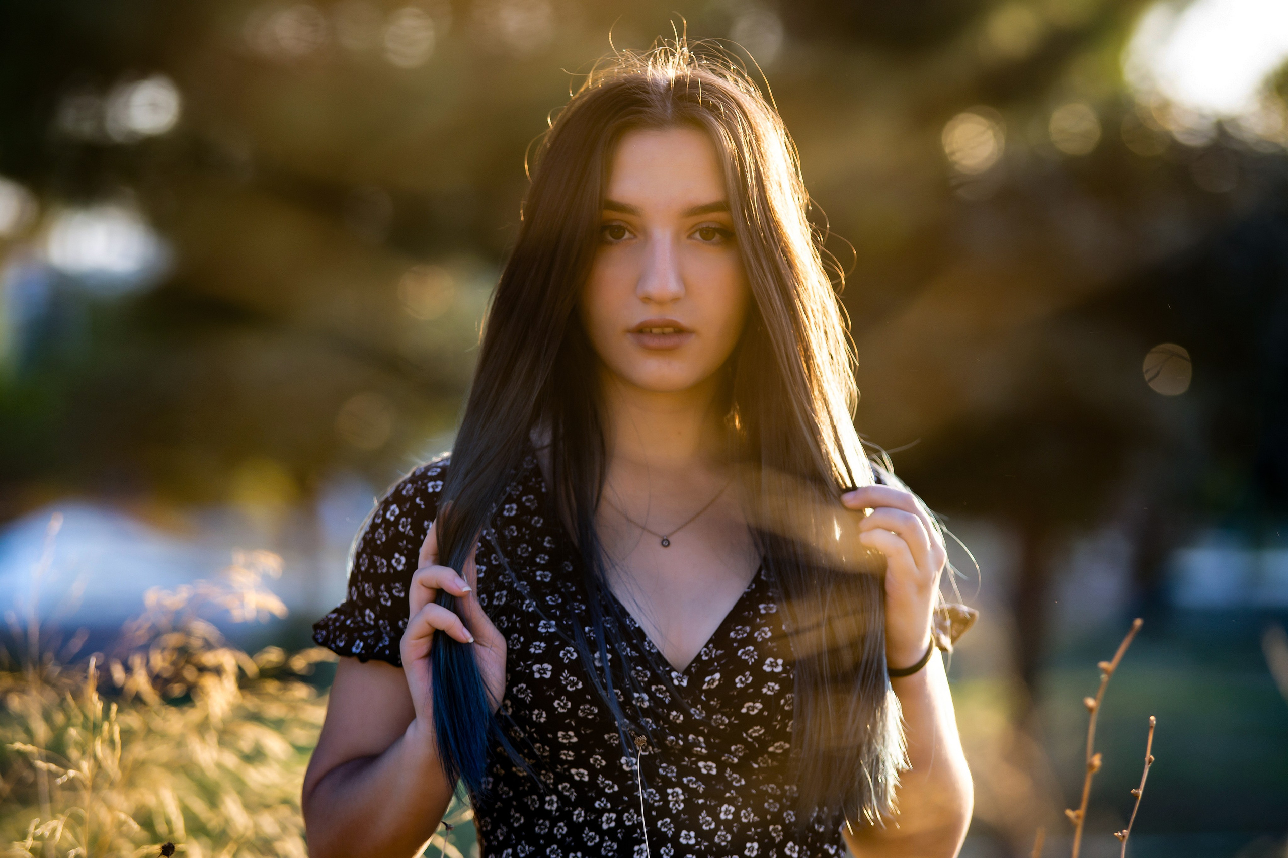 A young woman with long brown hair wearing a black floral print dress is posing outdoors in tall grass during golden hour, with sunlight shining through her hair and the background trees.