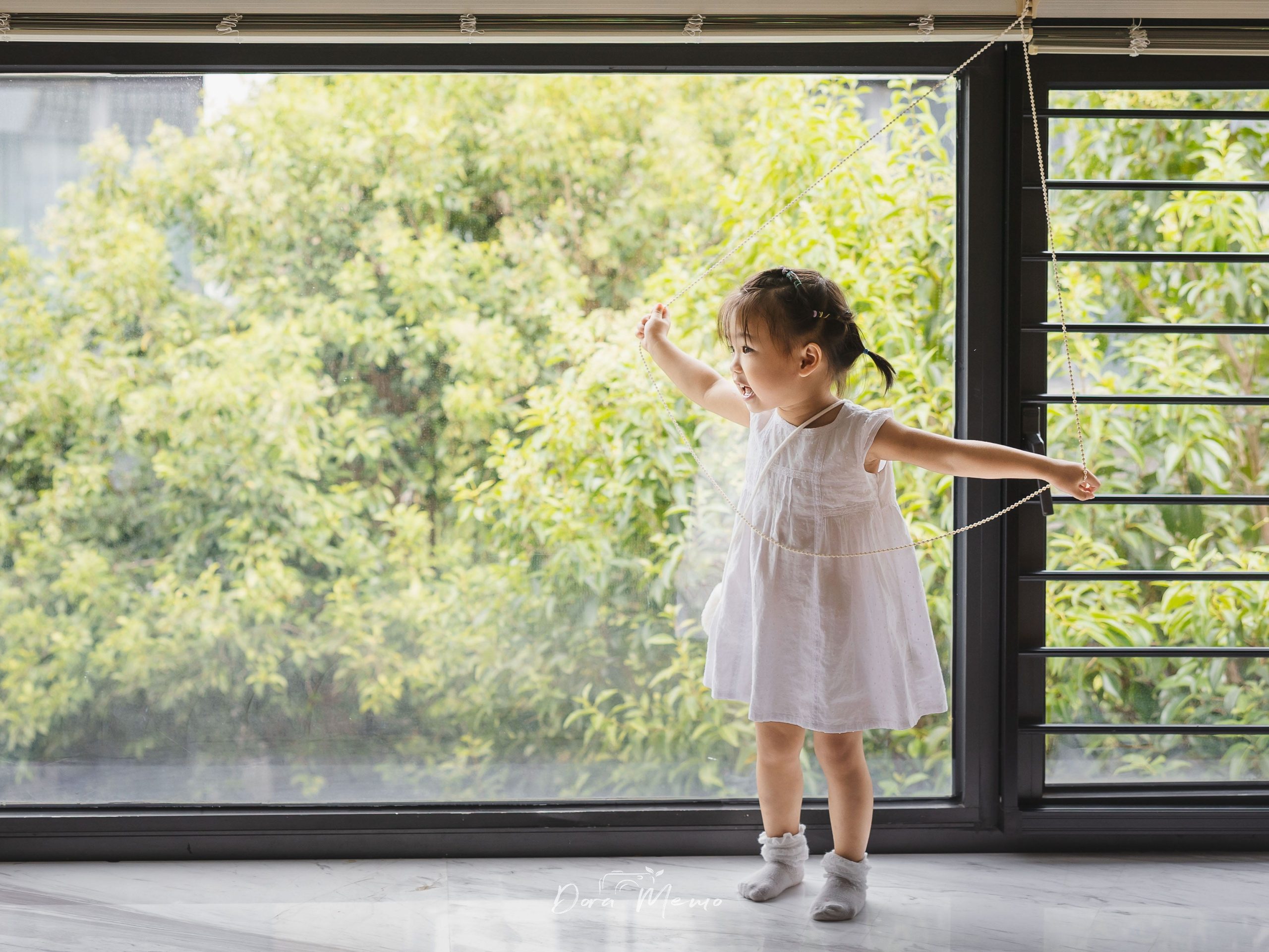 A young child stands beside a wide window, photographed in soft natural light at home.