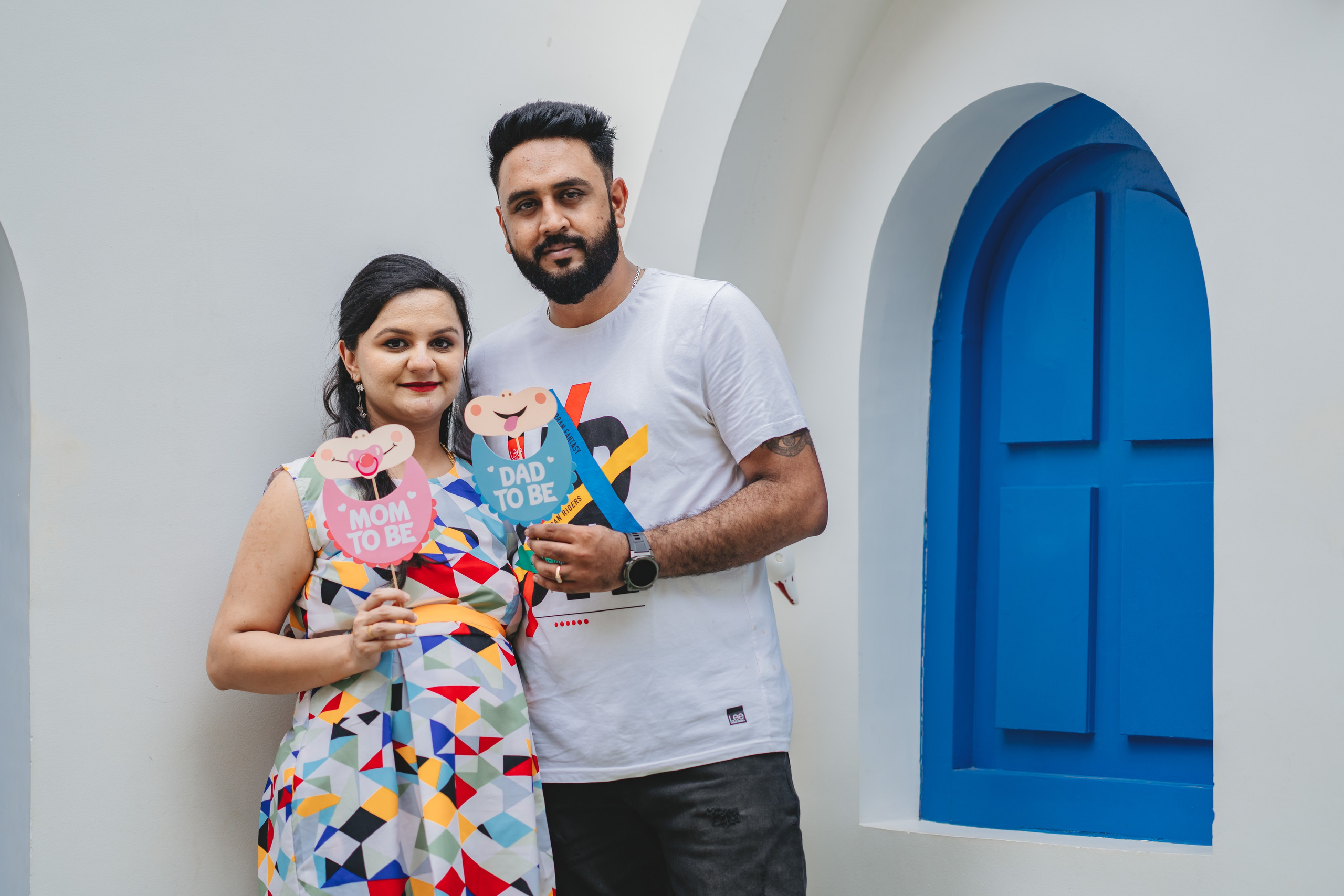 Maternity photoshoot in Bengaluru featuring a man and woman holding "Mom to Be" and "Dad to Be" props in front of a blue window backdrop.