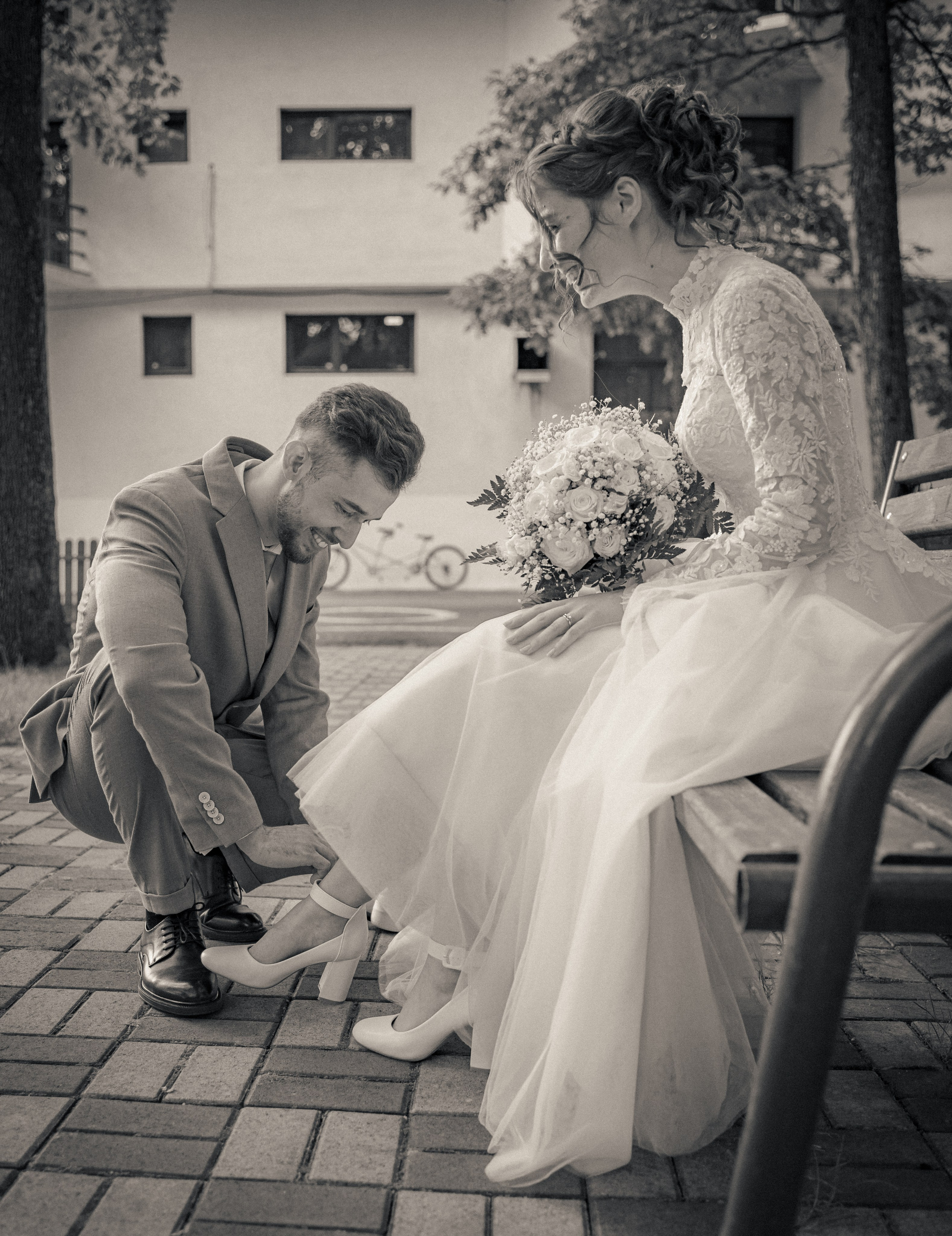 Fotografie alb-negru cu mirele aplecat să ajute mireasa cu pantofii pe o bancă, moment de grijă și complicitate, incadru.ro.