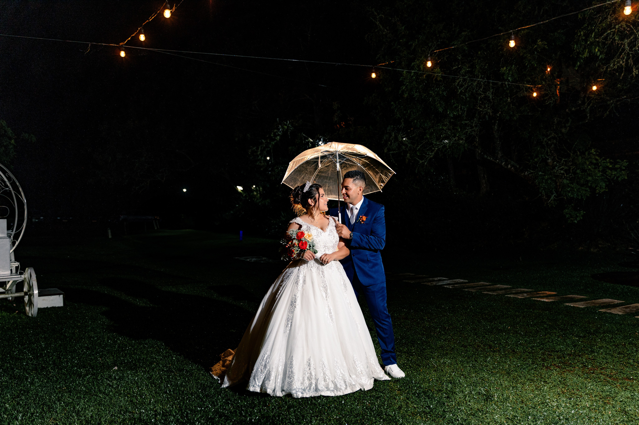 Noivos sob o guarda-chuva em área externa com luzes pendentes, composição romântica e criativa.