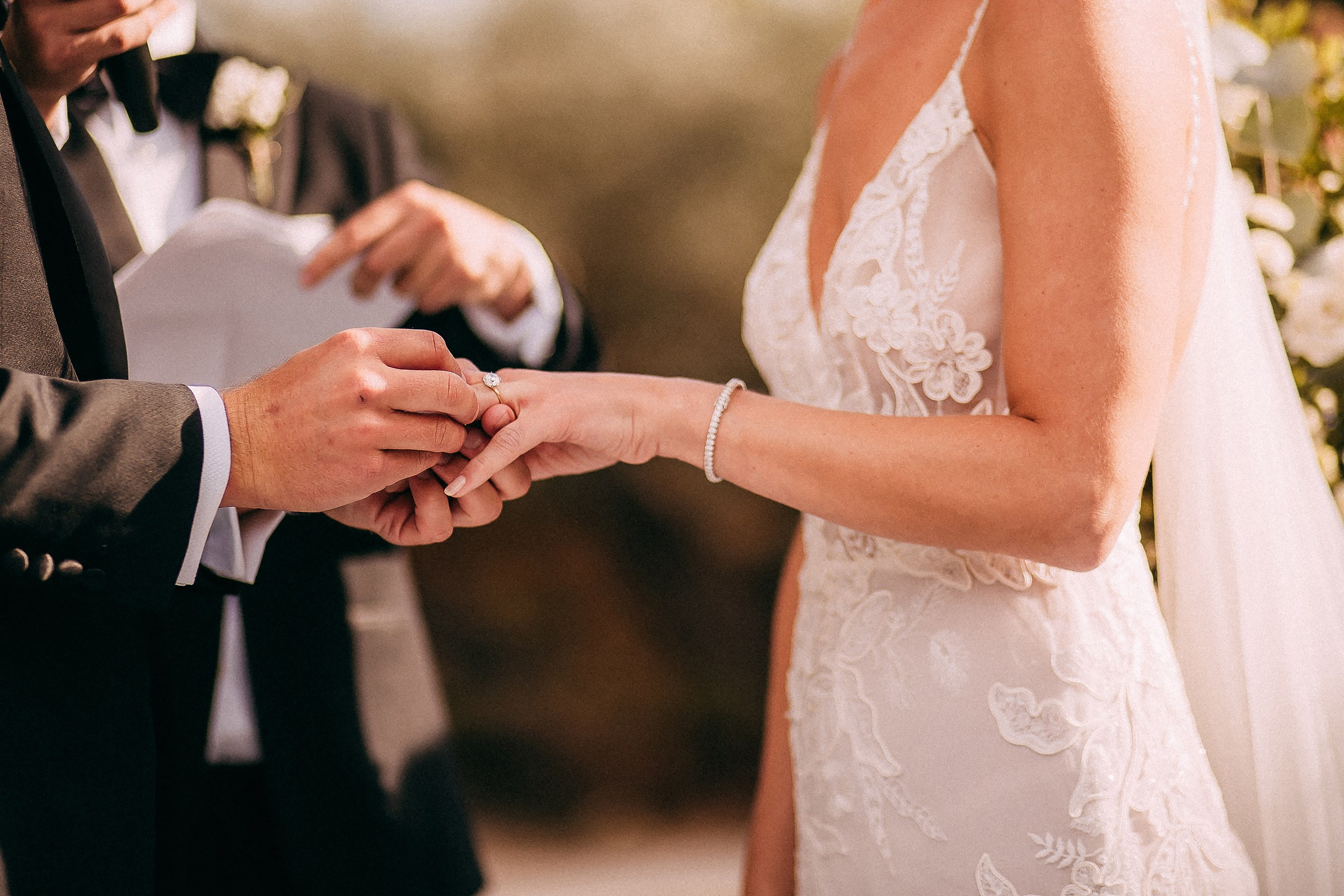 A close-up shot of a groom slipping a wedding band onto the bride’s finger, showcasing intricate details of her lace dress and delicate bracelet under soft sunlight.